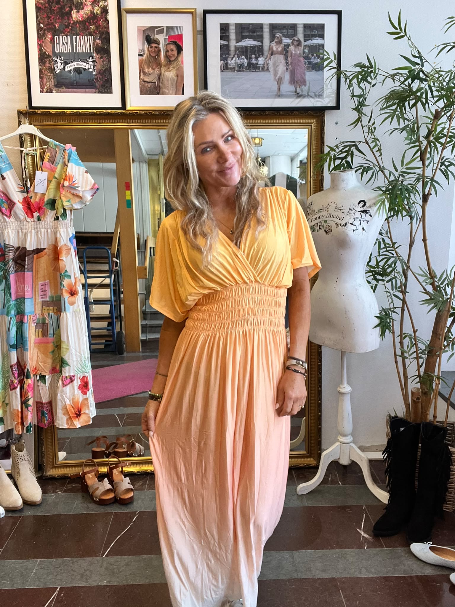 Leende kvinna i en lång persikofärgad klänning står och poserar i en butik. Runt henne syns en blommig klänning på galge, skor på golvet, provdocka med texten ”Fanny Michel” och inramade foton, inklusive ”CASA FANNY”.