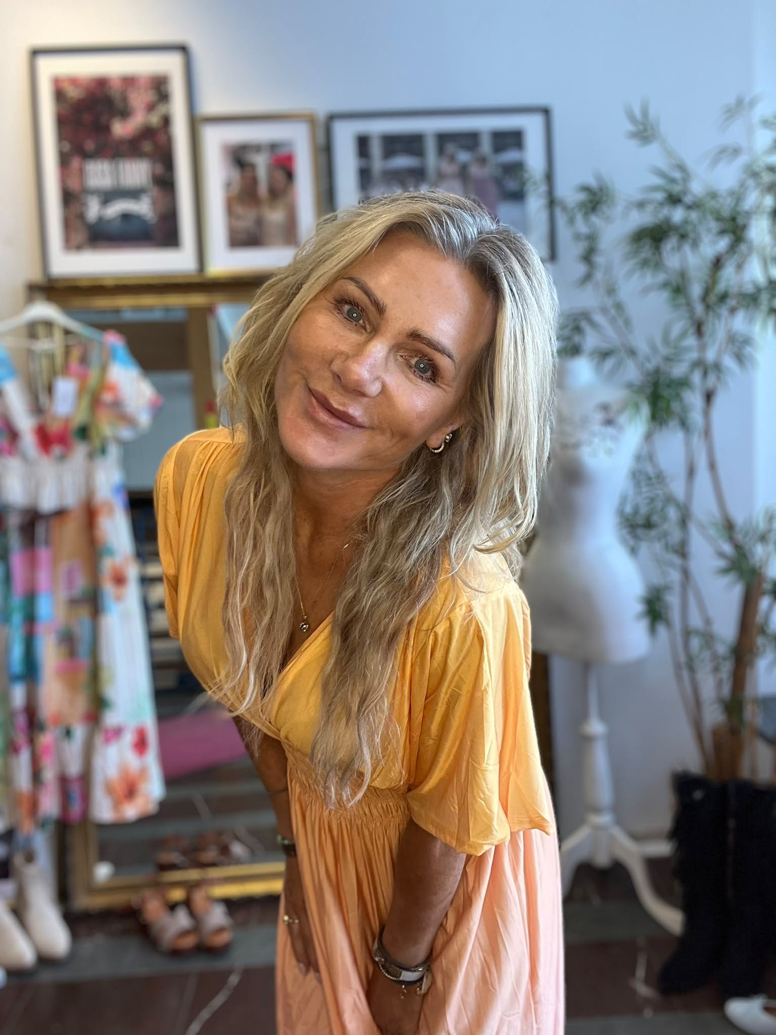 Leende blond kvinna i gul topp och persikofärgad kjol lutar sig fram mot kameran. Hon står i ett rum med klädställning, inramade foton på väggen och en stor krukväxt i bakgrunden.