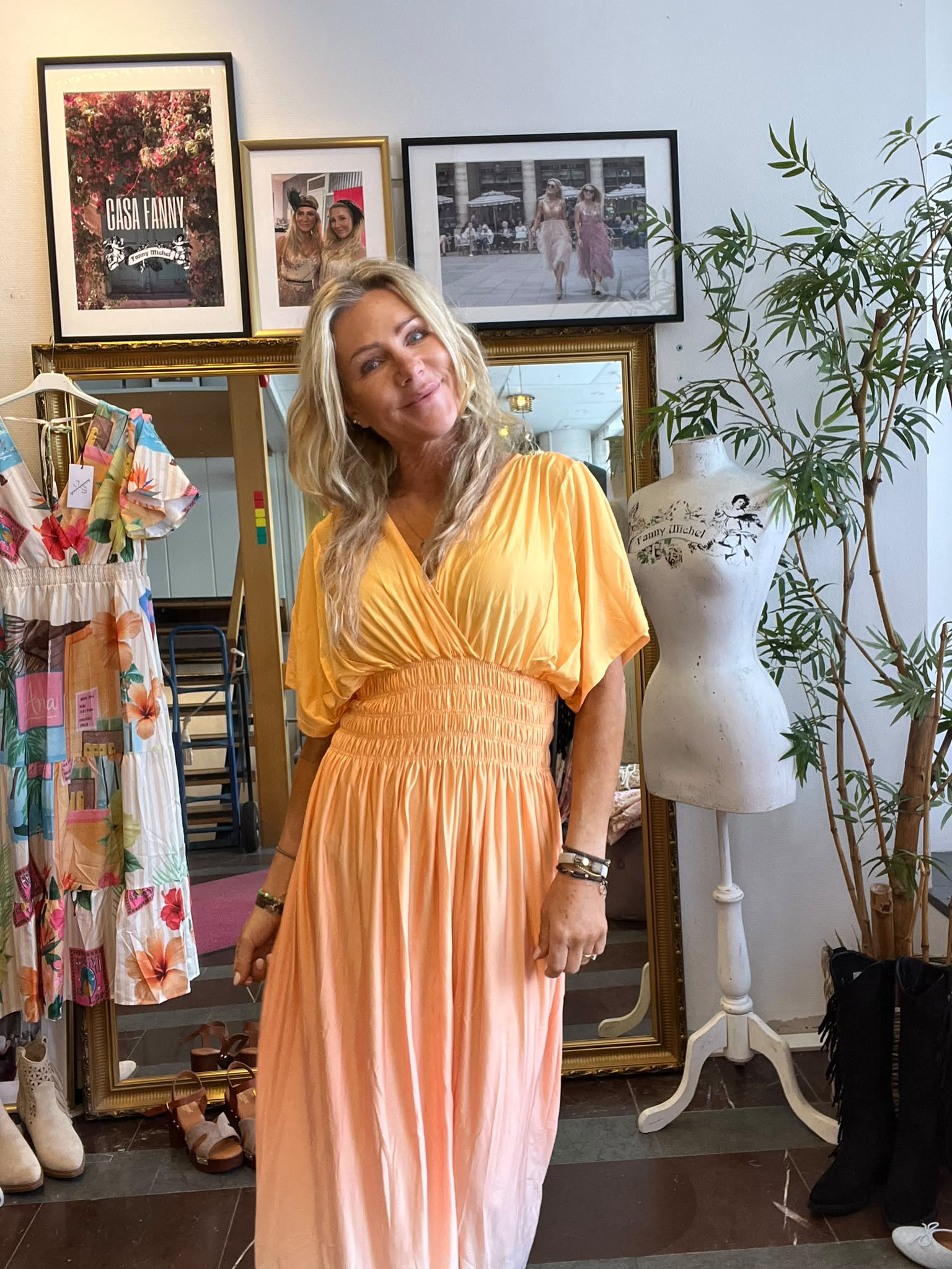Kvinna i lång orange klänning ler och poserar i en klädbutik, framför en stor guldramsspegel. Bredvid står en provdocka och en blommig klänning på galge; på väggen syns ramar med texten “CASA FANNY”.