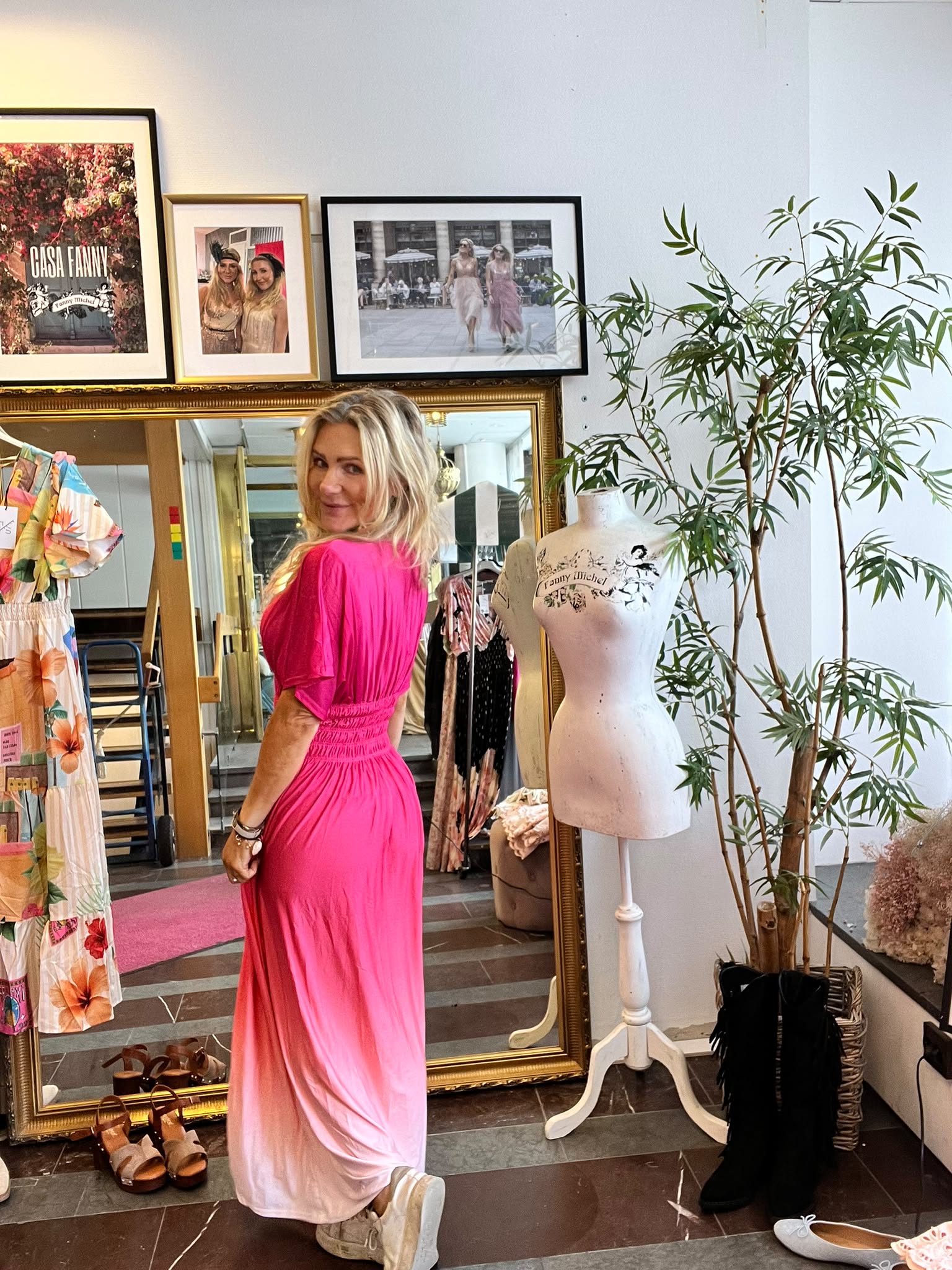Blond kvinna i lång rosa klänning poserar och ser över axeln i en klädbutik, framför en stor guldspegel. Runt henne syns klänningar och skor, provdocka med texten “family titchel” samt tavla med “CASA FANNY”.