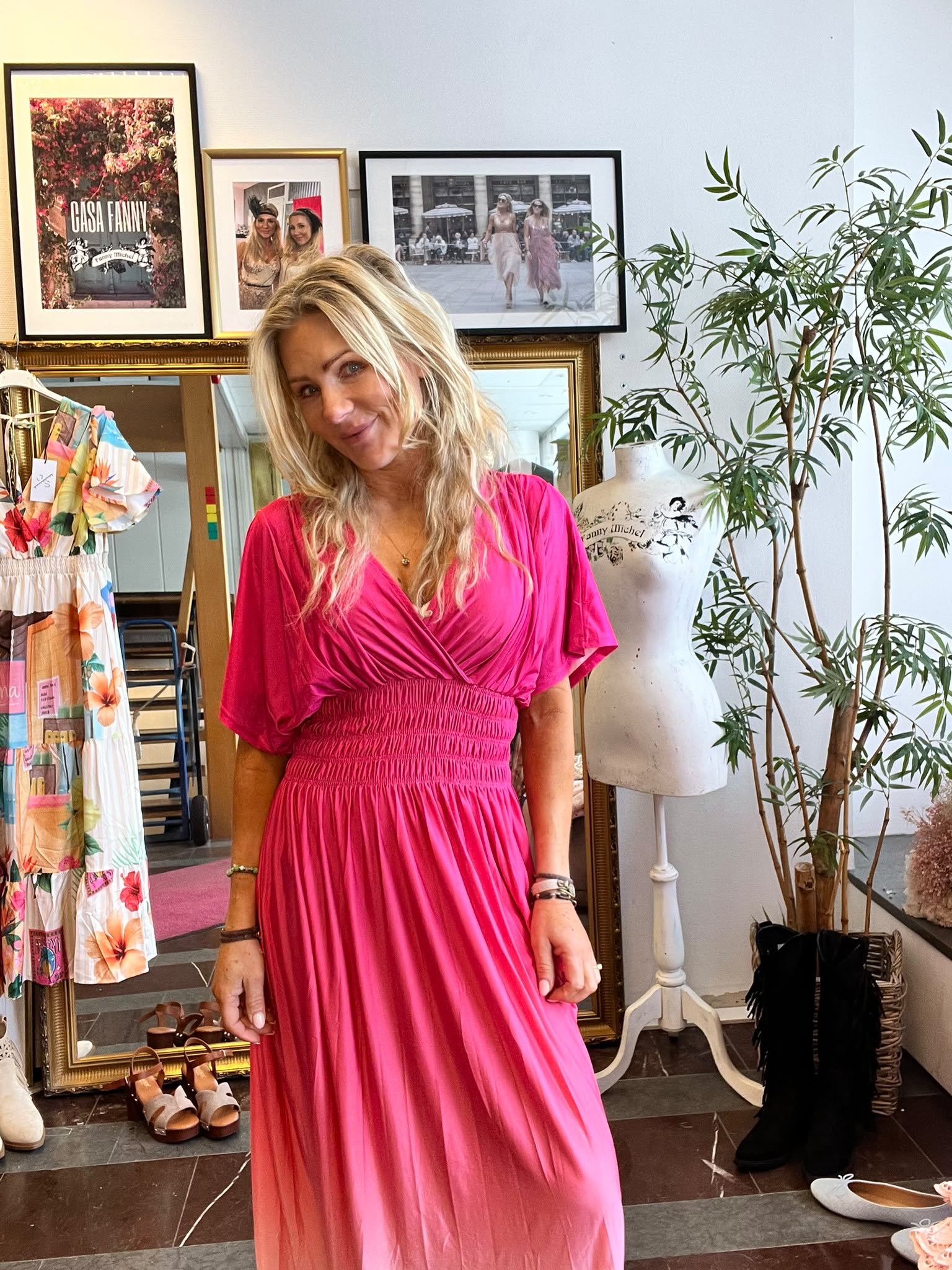 Leende kvinna i ceriserosa omlottklänning poserar i en klädbutik/studio, framför en spegel och inramade foton. Till höger syns en provdocka och en stor grön växt, till vänster hänger en färgstark klänning. Text på tavla: ”CASA FANNY”.
