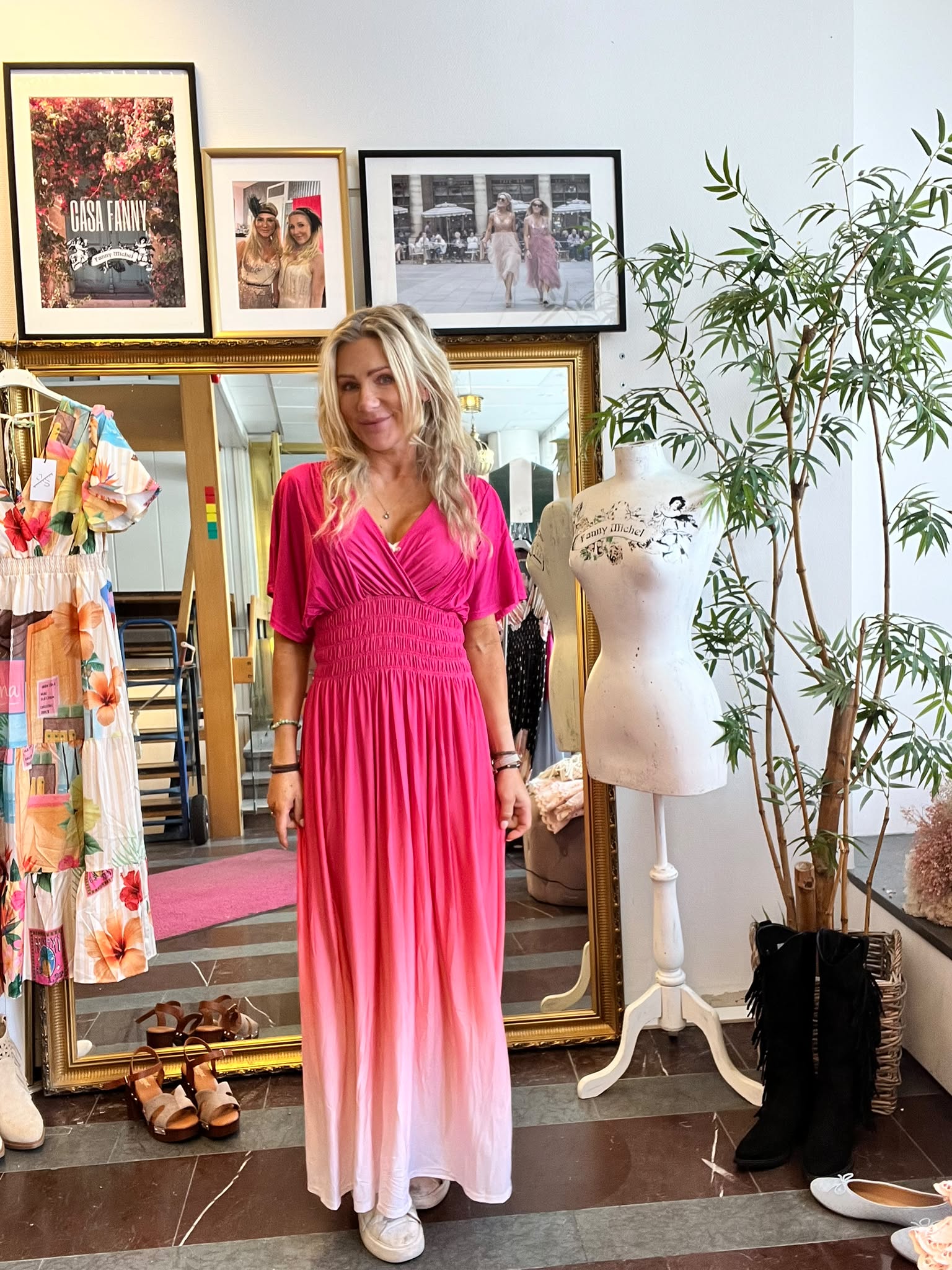 Blond kvinna står och ler i en klädbutik, iklädd en rosa maxiklänning med ombre mot vitt. Runt henne syns spegel, klänningar och skor, provdocka med texten ”Fanny Michel”, samt tavla med texten ”CASA FANNY”.