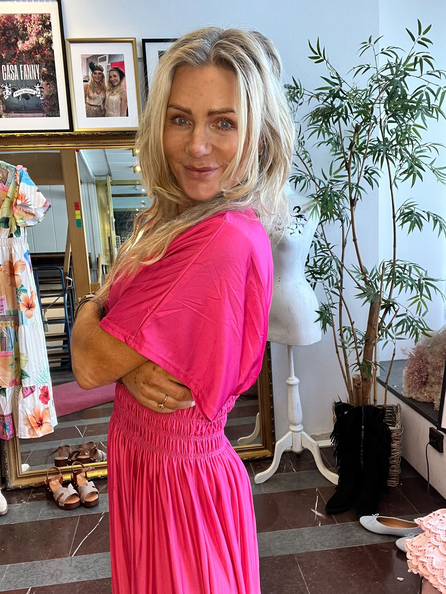 Blond kvinna i starkt rosa klänning står med armarna i kors och ler mot kameran i en butik/showroom. Bakom syns stor guldspegel, kläder och skor samt inramade foton med texten ”CASA FANNY”.