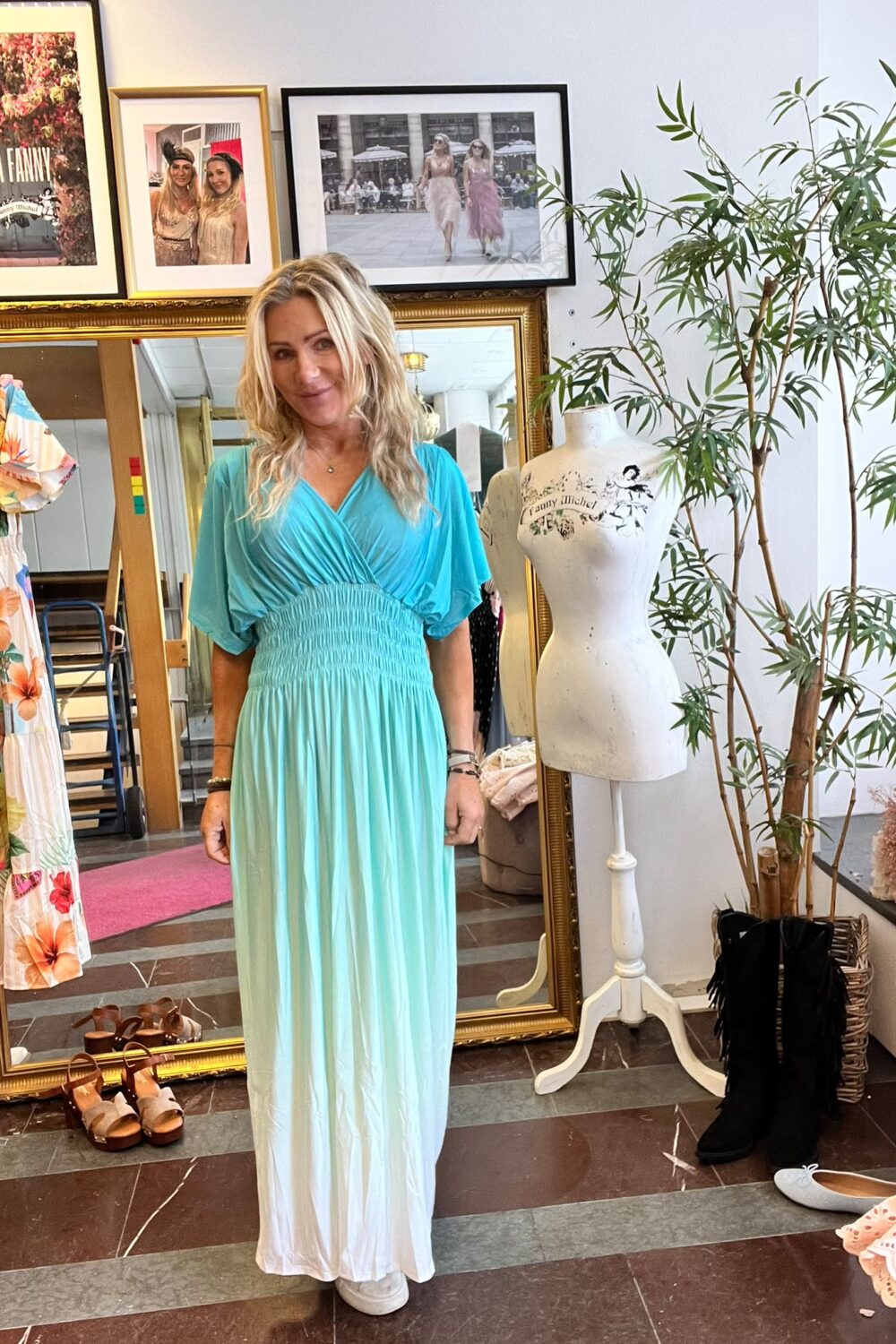 Blond kvinna står och ler i en butik, klädd i turkos omlottklänning med lång vit nederdel. Bredvid finns en provdocka och en stor spegel; på väggen hänger ramar med texten ”CASA FANNY” och ”Fanny Michel”.