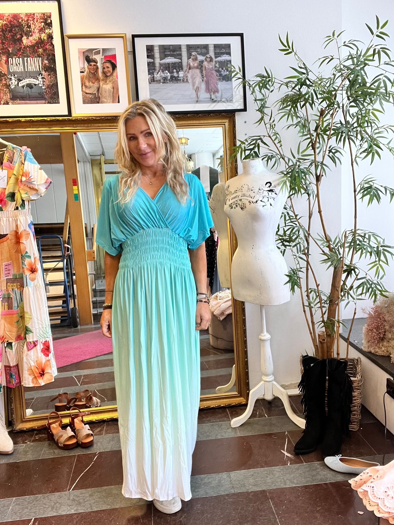 Blond kvinna står och ler i en butik, klädd i turkos omlottklänning med lång vit nederdel. Bredvid finns en provdocka och en stor spegel; på väggen hänger ramar med texten ”CASA FANNY” och ”Fanny Michel”.