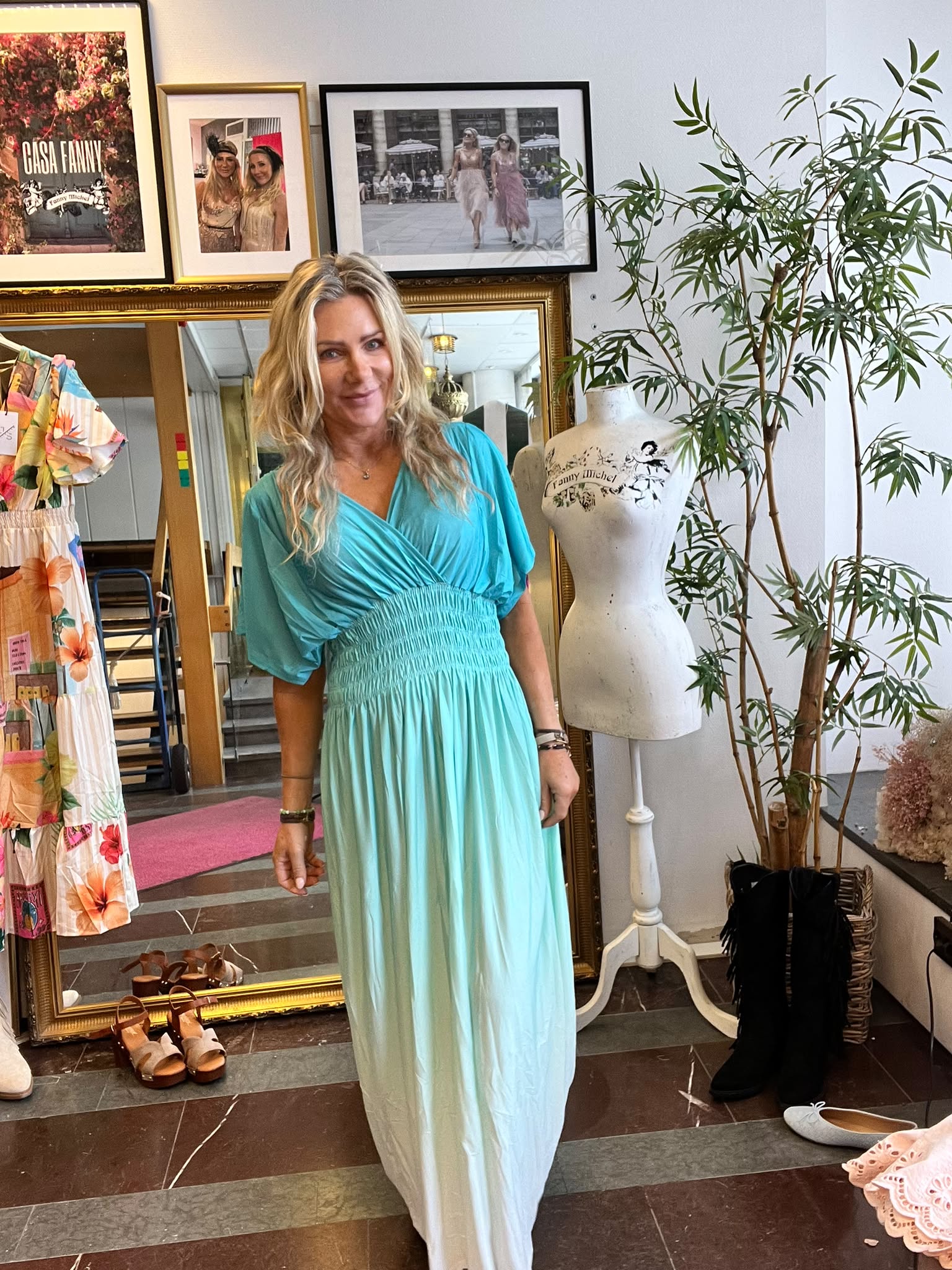 Kvinna i turkos klänning står och ler i en klädbutik, framför en stor guldspegel. Bredvid syns en skyltdocka med texten “Fanny Michel” och på väggen en affisch med texten “CASA FANNY”.