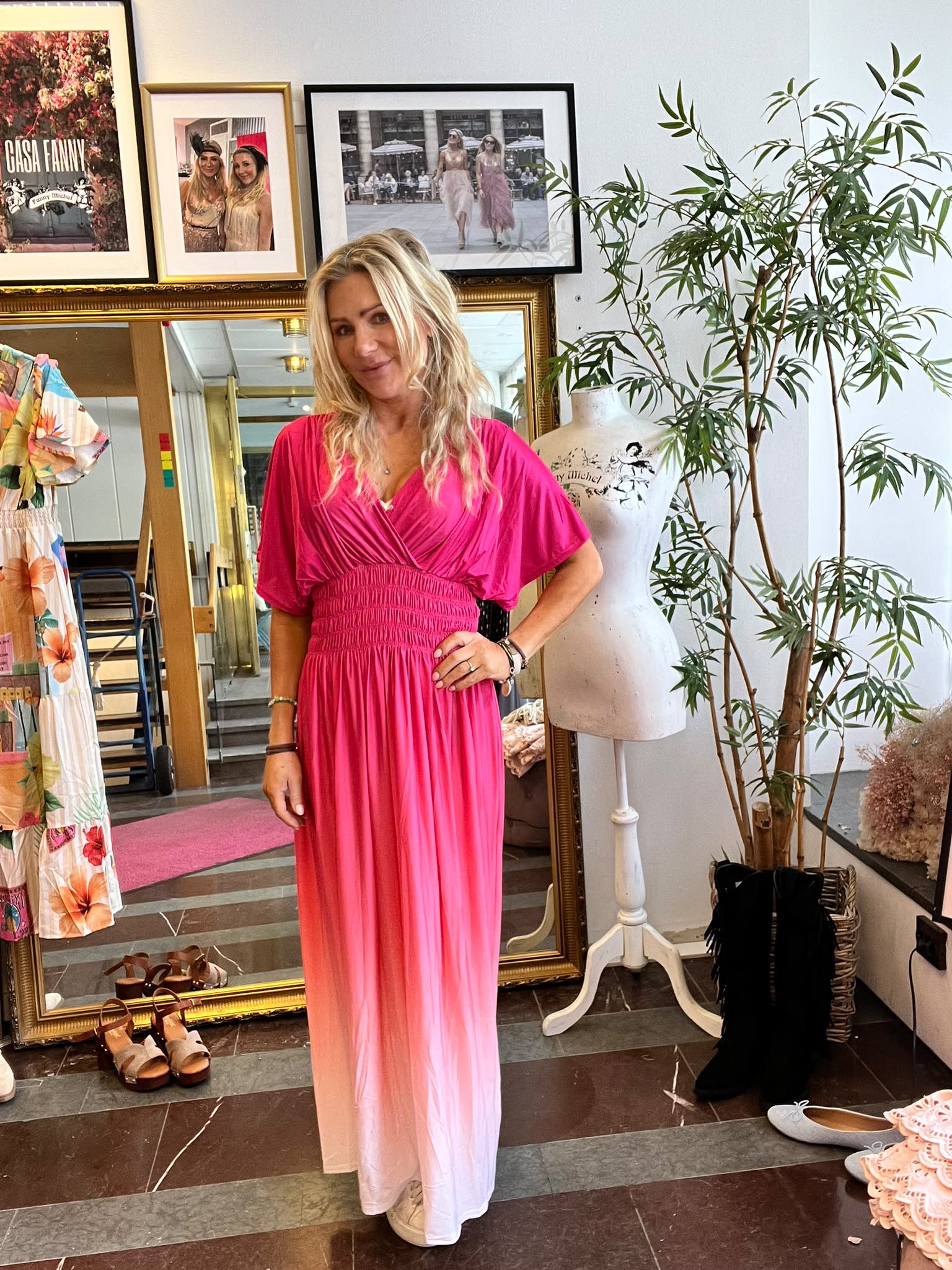 Blond kvinna poserar med handen på höften i en starkt rosa, lång klänning i en butik. Runt henne syns en stor guldspegel, klänningar på galge, skor på golvet, provdocka och en hög grön växt. Text: “CASA FANNY”.