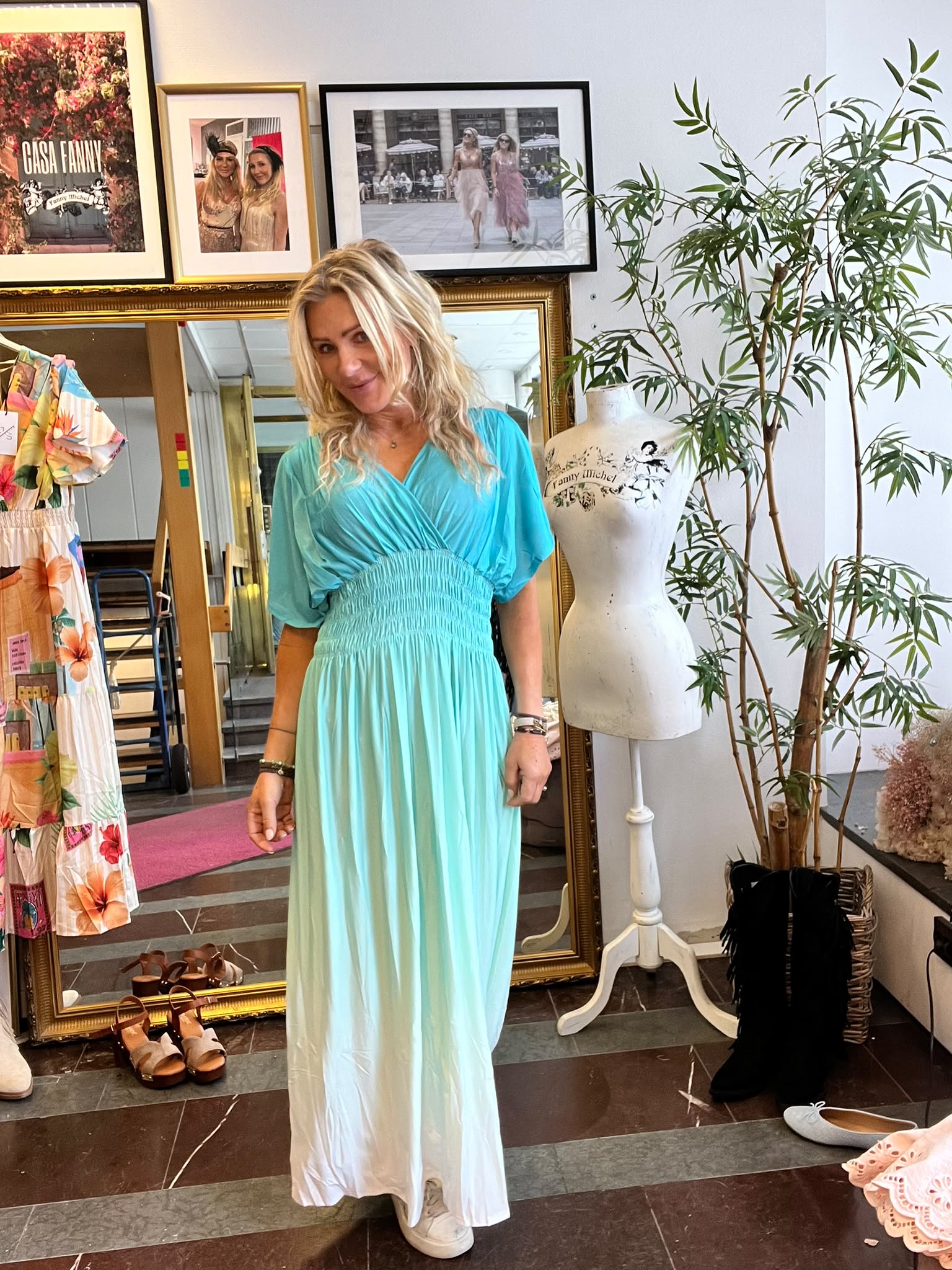 Blond kvinna står och ler i en butik/ateljé, iklädd turkos maxiklänning och vita sneakers. Runt henne syns spegel, klädställning och skor samt provdocka märkt ”Fanny Michel” och tavla med texten ”CASA FANNY”.
