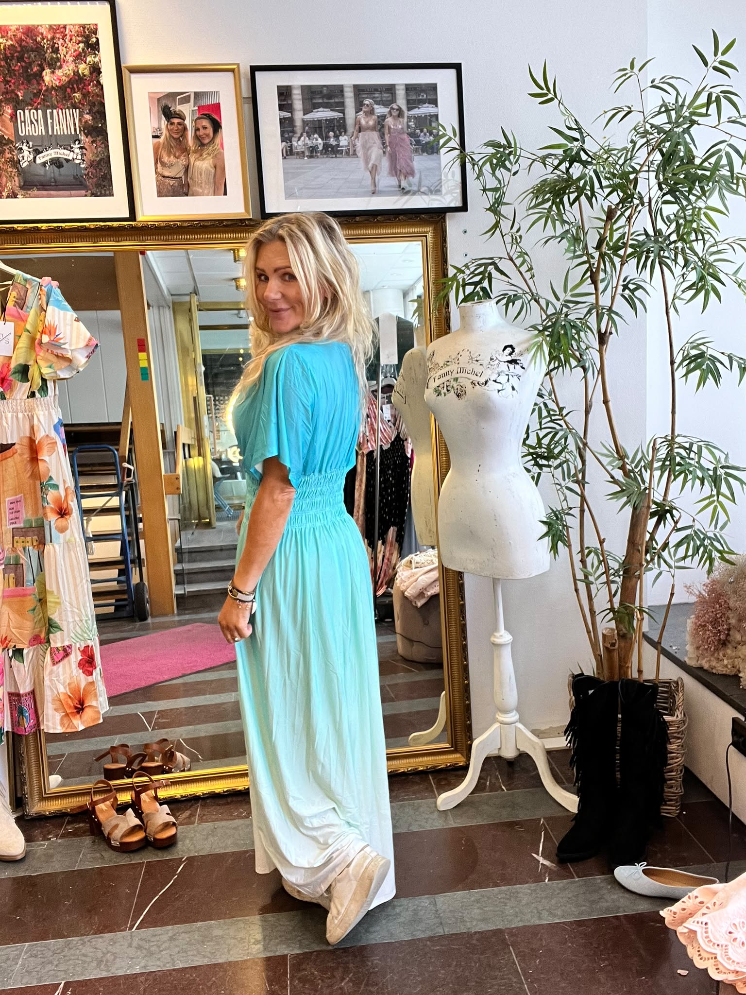 Blond kvinna provar en turkos klänning och tittar leende över axeln framför en stor guldramsspegel i en butik. Runt henne syns skor på golvet, klänningar på galge och ett provdockshuvud med texten “Fanny Michel”.