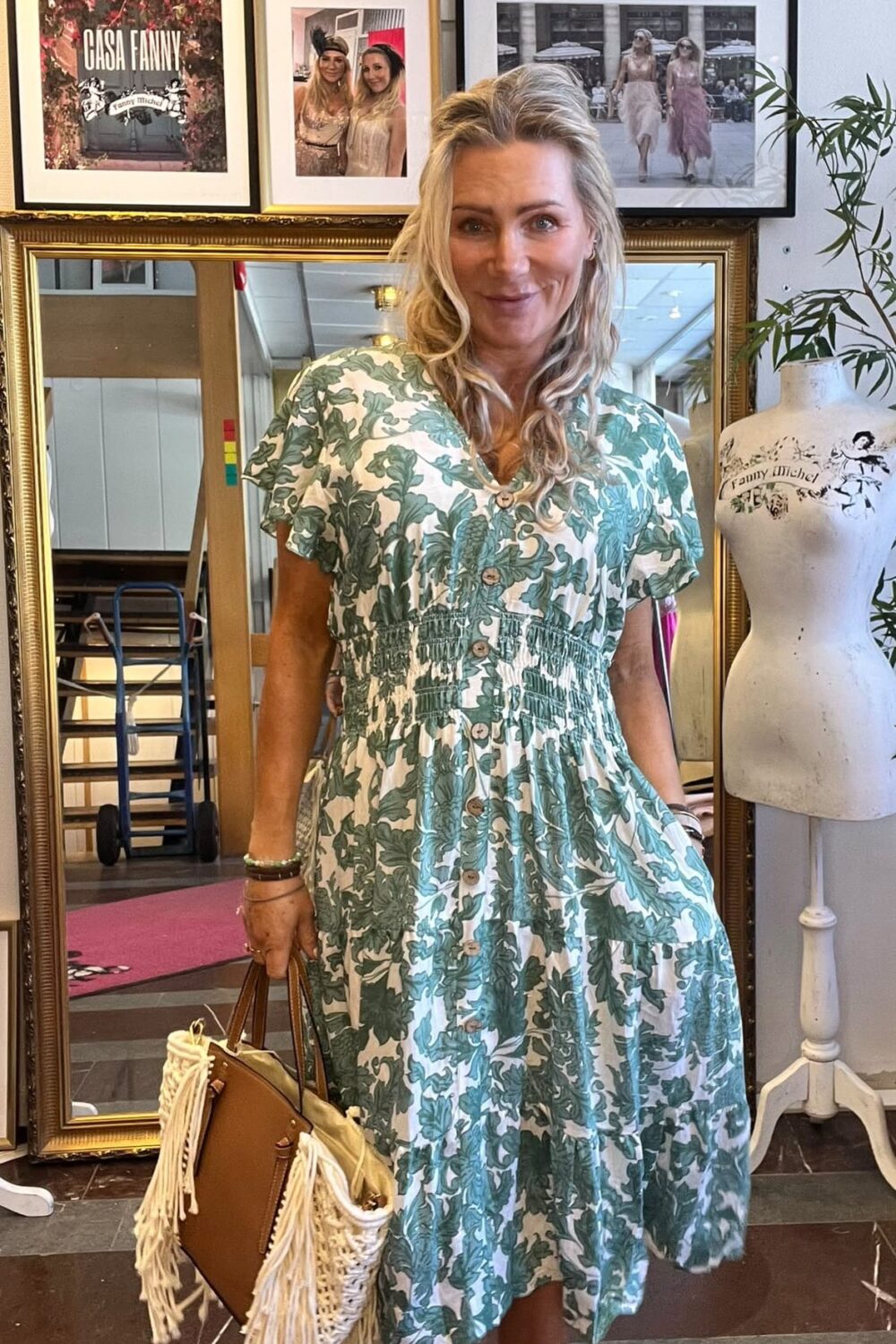 Kvinna i grönvitt mönstrad klänning ler mot kameran och håller en brun handväska med fransar i en butik. Bakom syns spegel, provdocka och inramade foton med texten “CASA FANNY” och “Fanny Michel”.