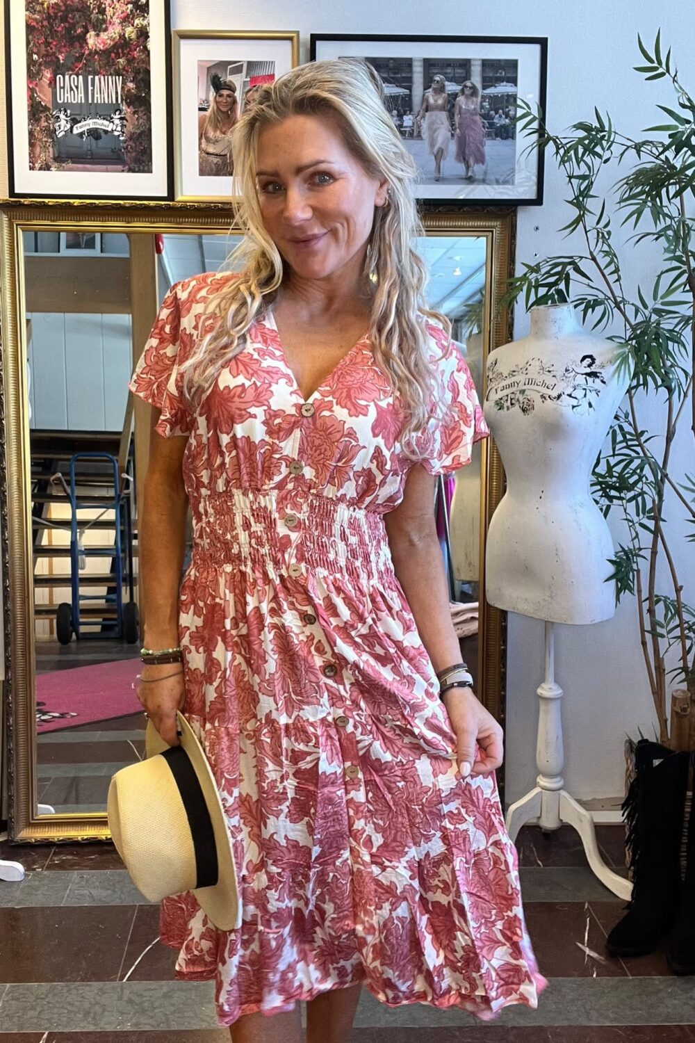 Leende kvinna i rosa‑vit blommig klänning håller en stråhatt och poserar inomhus framför en stor guldram. På väggen bakom syns inramade foton med texten “CASA FANNY” och en provdocka märkt “Fanny Michel”.