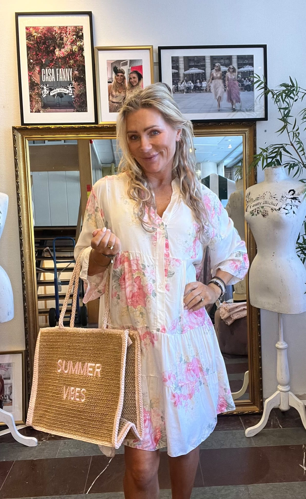 Leende kvinna i blommig, ljus klänning poserar och håller en stråväska med texten ”SUMMER VIBES”. Hon står framför en stor spegel och inramade bilder på väggen, bredvid en provdocka i en butik. Text i tavla: ”GISA FANNY”.