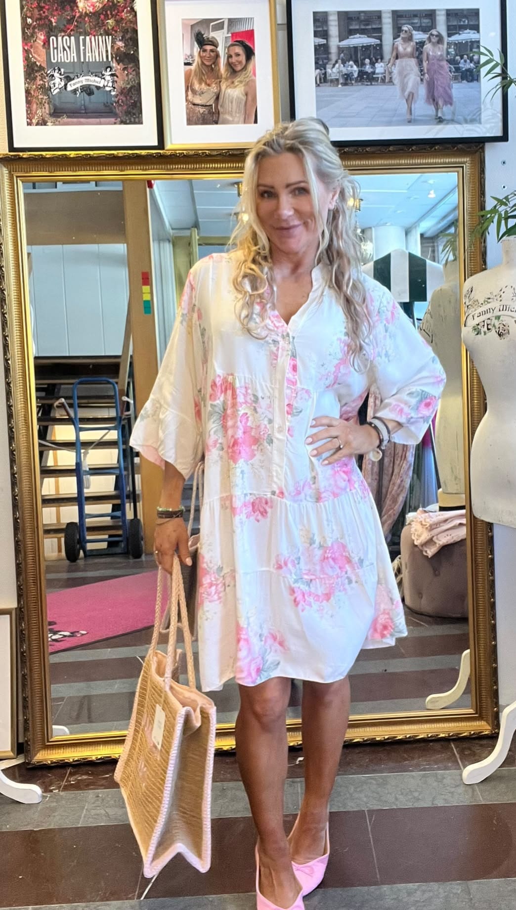 Blond kvinna poserar framför en stor guldramsspegel, med handen på höften och en flätad strandväska i andra handen. Hon bär vit klänning med rosa blommor och rosa klackar, i en butik med inramade foton och texten ”CASA FANNY”.
