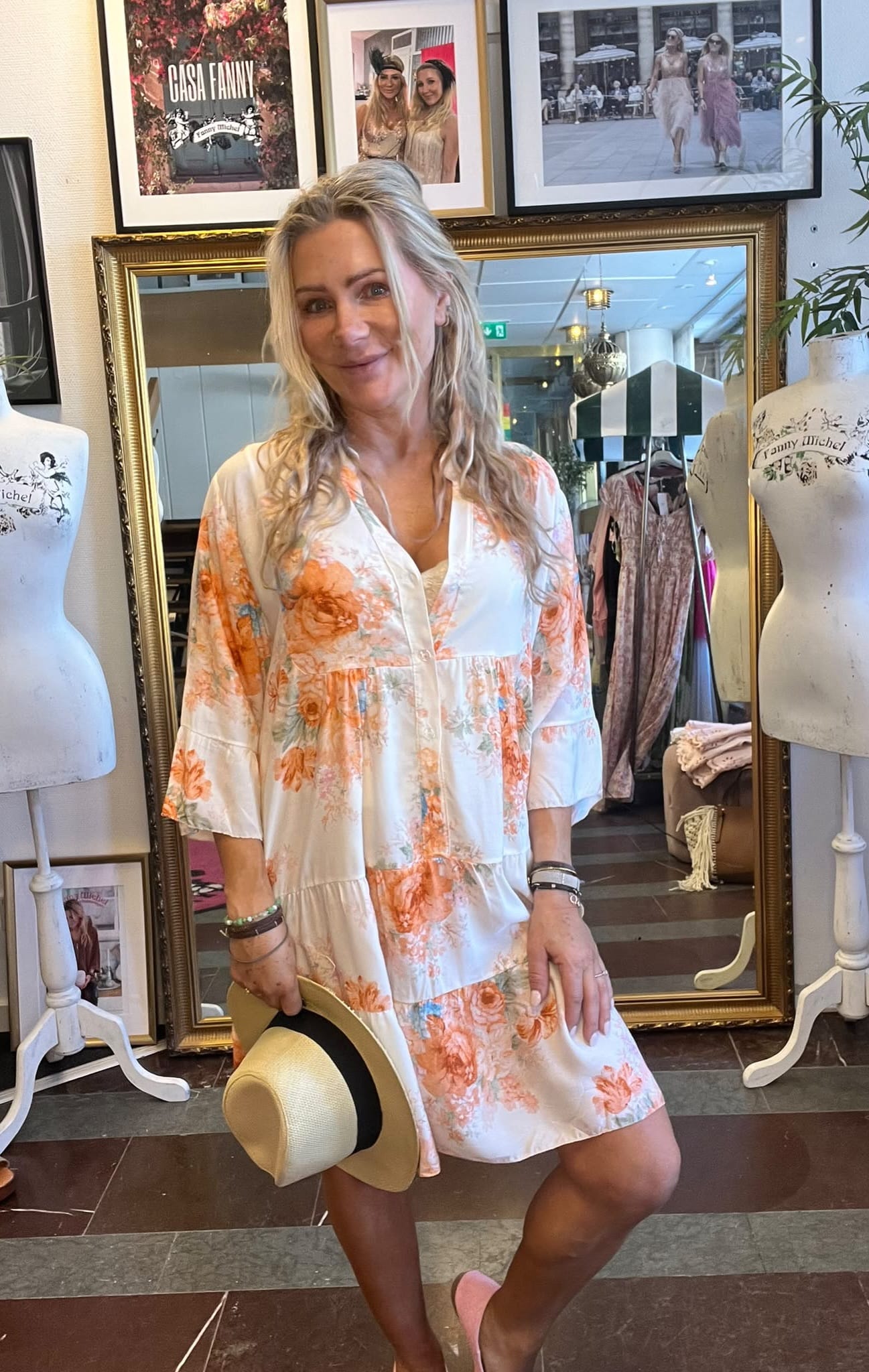 Leende kvinna står framför en stor guldramsspegel i en klädbutik, klädd i vit, blommig klänning och håller en stråhatt. Runt henne syns provdockor och inramade foton med texten ”CASA FANNY” och ”Fanny Michel”.
