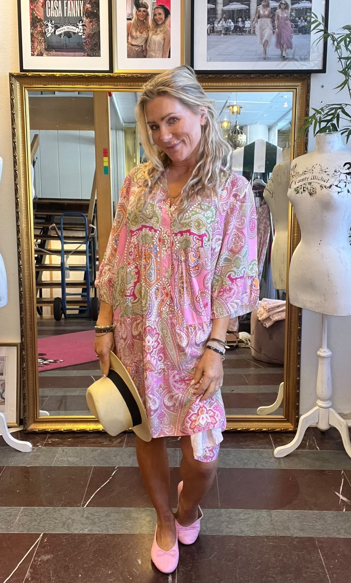Blond kvinna står och ler framför en stor guldramsspegel, håller en ljus hatt med svart band och bär rosa mönstrad tunika och rosa skor. I bakgrunden syns provdocka med texten ”Fanny Michel” samt skylt ”CASA FANNY”.
