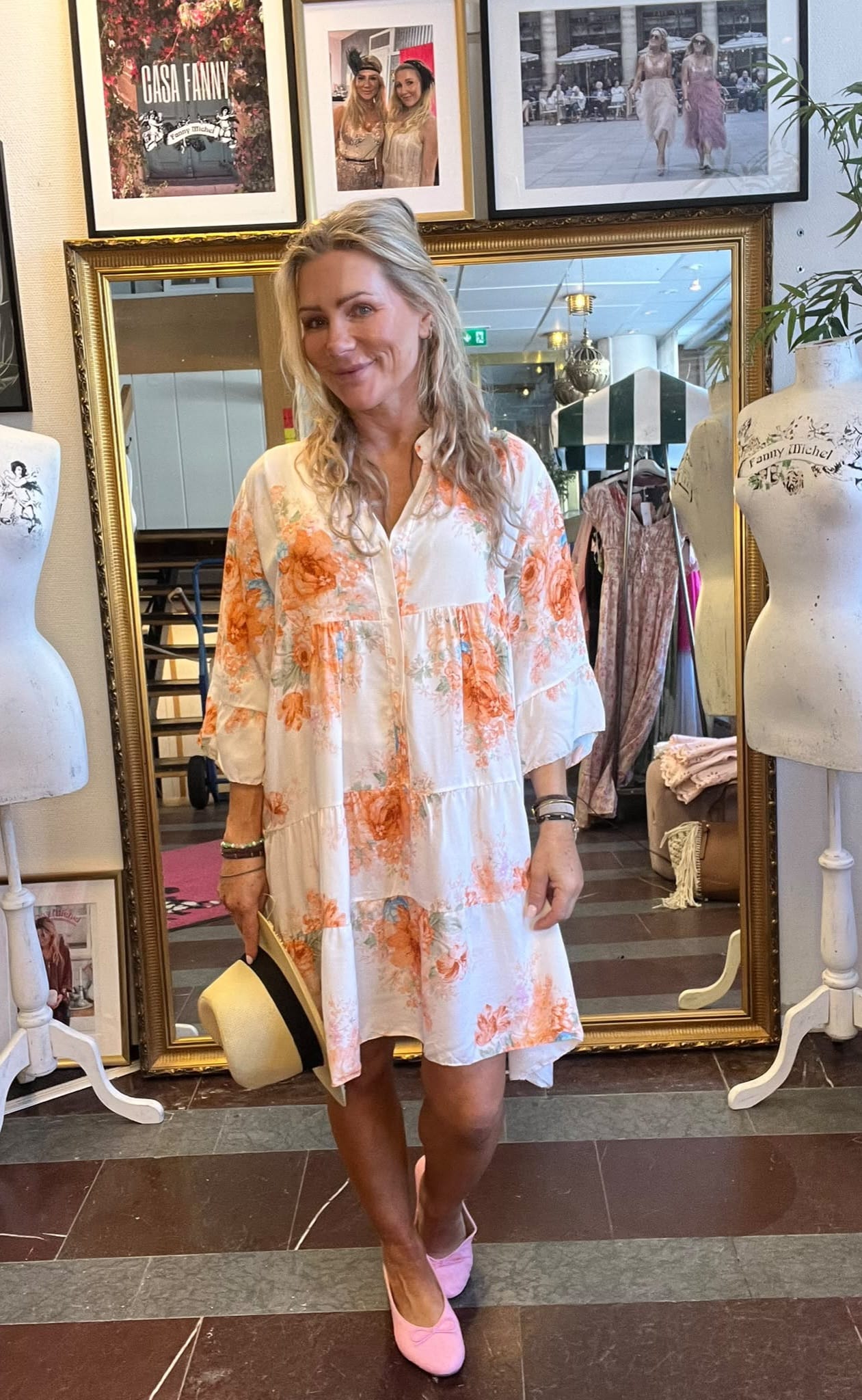 Leende kvinna står framför en stor guldramsspegel i en klädbutik, bär vit klänning med orange blommor och håller en ljus hatt. På väggen bakom syns inramade foton och texten ”CASA FANNY”.
