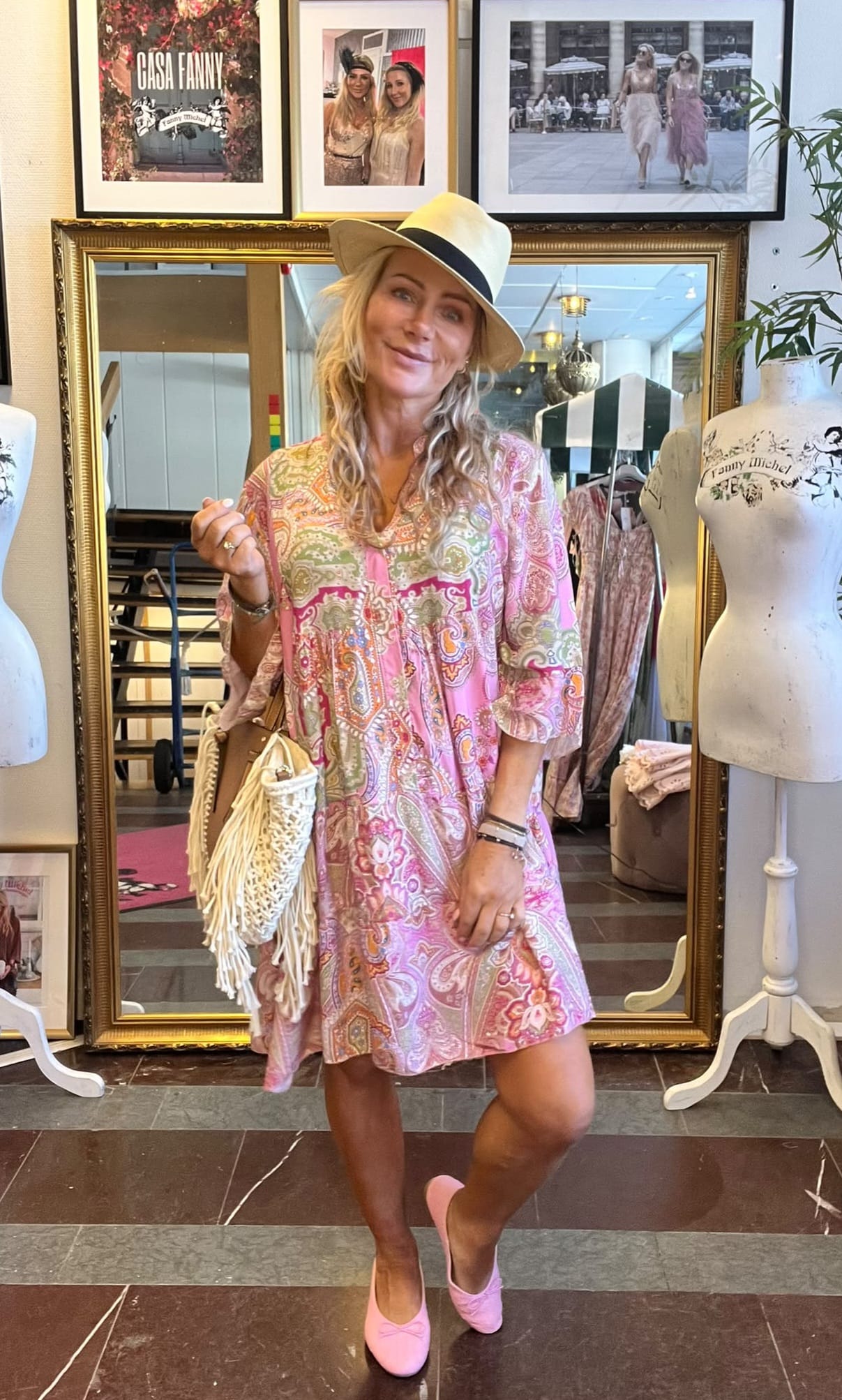 Kvinna i beige hatt poserar och håller en fransad vit väska i en klädbutik, iklädd rosa mönstrad klänning och rosa skor. Bakom syns en stor spegel, provdockor och inramade foton med texten ”CASA FANNY”.
