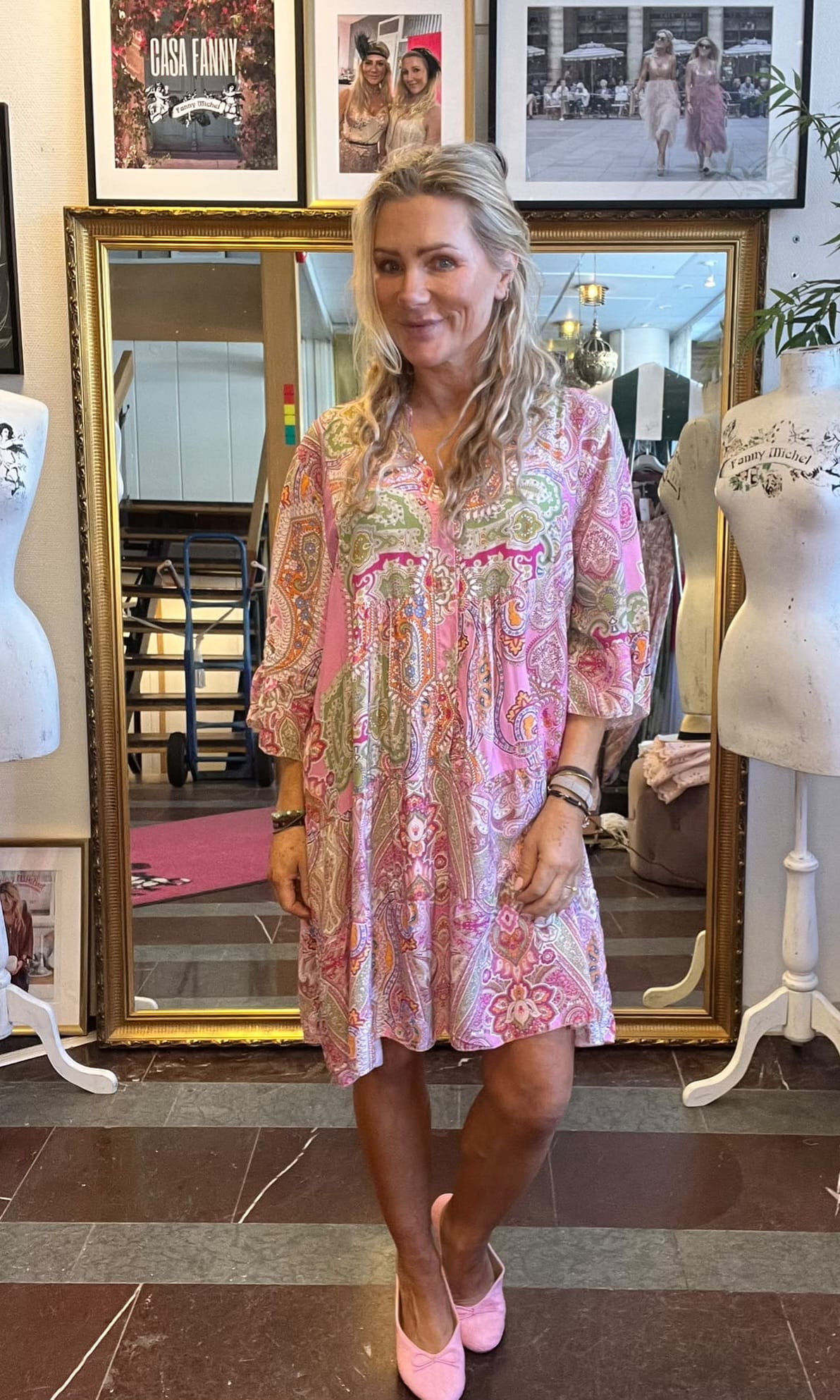 Kvinna står och ler framför en stor guldramsspegel i en klädbutik, klädd i kort rosa mönstrad klänning och rosa skor. Runt henne syns provdockor och inramade foton; en tavla har texten “CASA FANNY”.