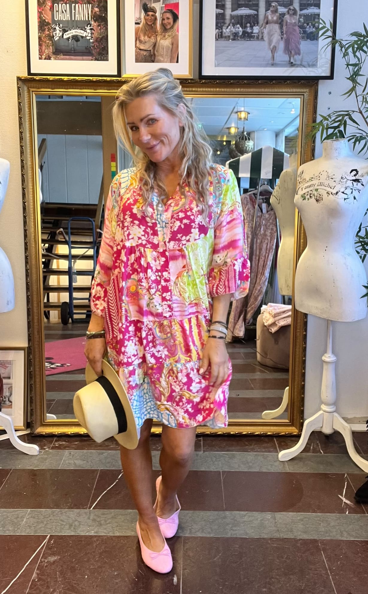 Kvinna poserar och ler i en färgstark, blommig klänning och håller en ljus hatt med svart band. Hon står i en butik framför en stor guldramsspegel, bredvid provdocka med texten “Fanny Michel”.