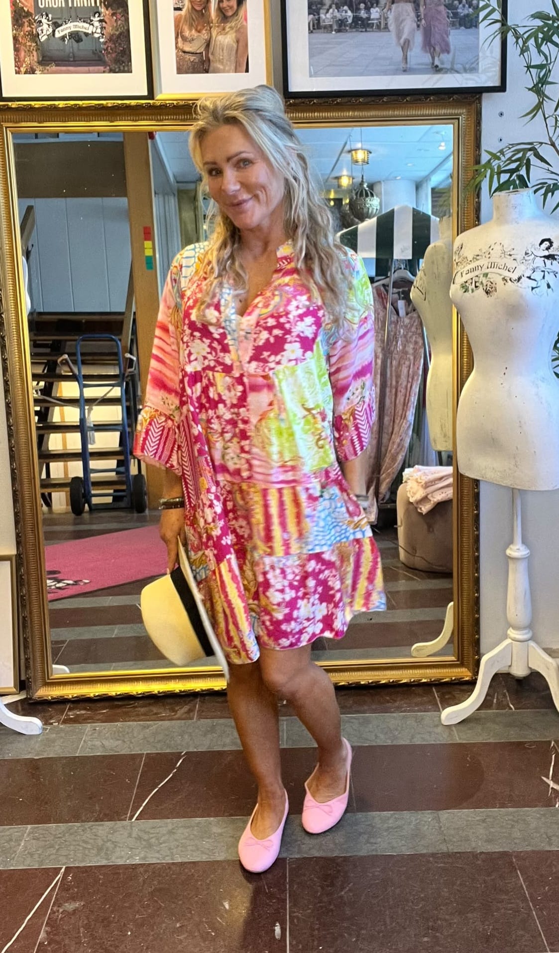 Kvinna poserar och ler i en butik, klädd i färgstark mönstrad klänning och rosa skor, hållande en ljus hatt. Bakom henne står en stor guldramsspegel, en provdocka och inramade foton på väggen.