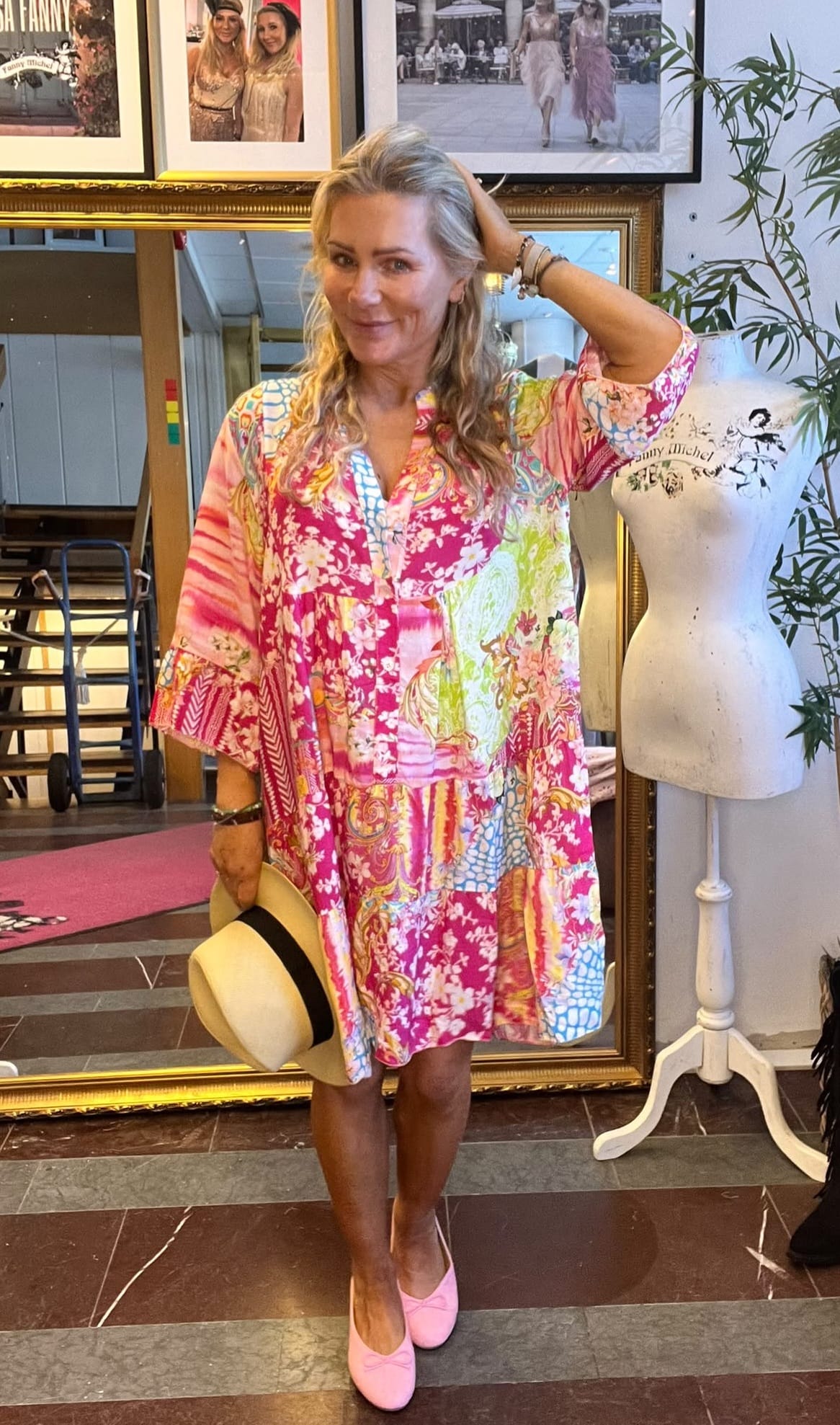 Blond kvinna poserar med ena handen i håret och håller en stråhatt. Hon bär en färgstark, blommig klänning och rosa skor, stående i en ateljé med speglar, inramade foton och en provdocka. Text på provdockan: ”Fanny Michel”.