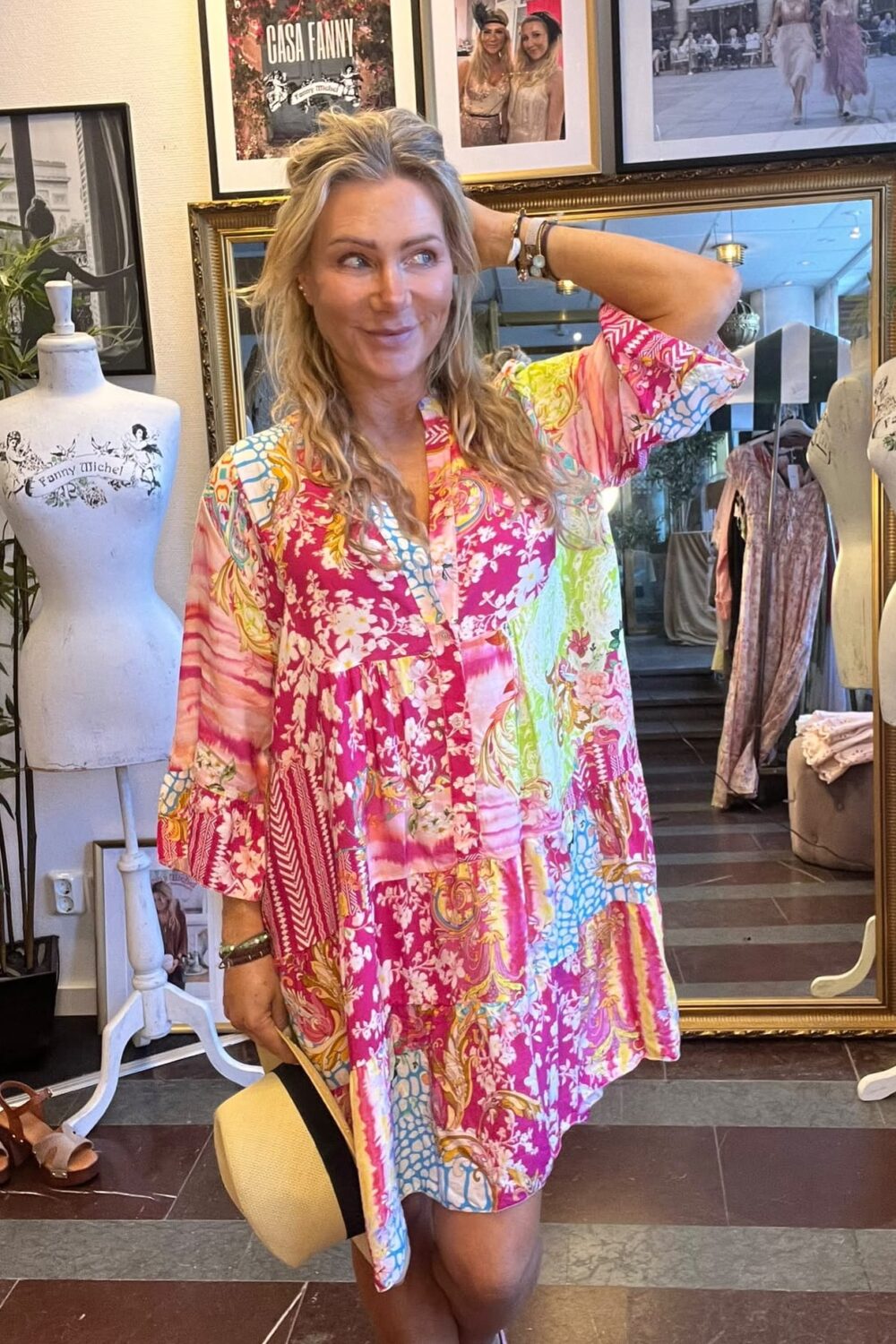 Kvinna i färgstark, blommig tunika står och ler med ena handen bakom huvudet, hållande en stråhatt. Hon är i en klädbutik med provdockor och spegel; på väggen syns texten “CASA FANNY”.