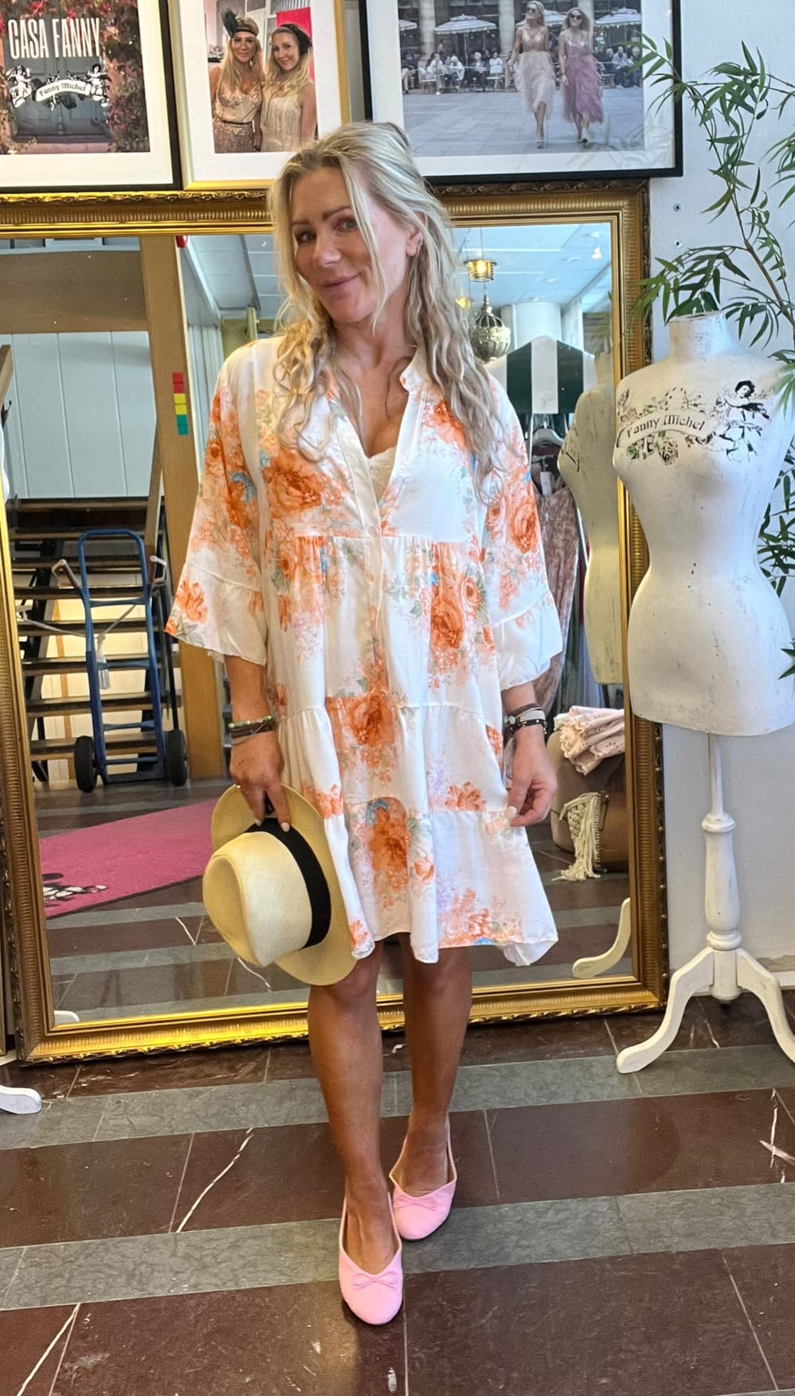 Leende kvinna står framför en stor guldramsspegel och håller en stråhatt med svart band. Hon bär vit, blommig klänning och rosa skor i en butik med provdocka märkt “Fanny Michel” och inramade foton.