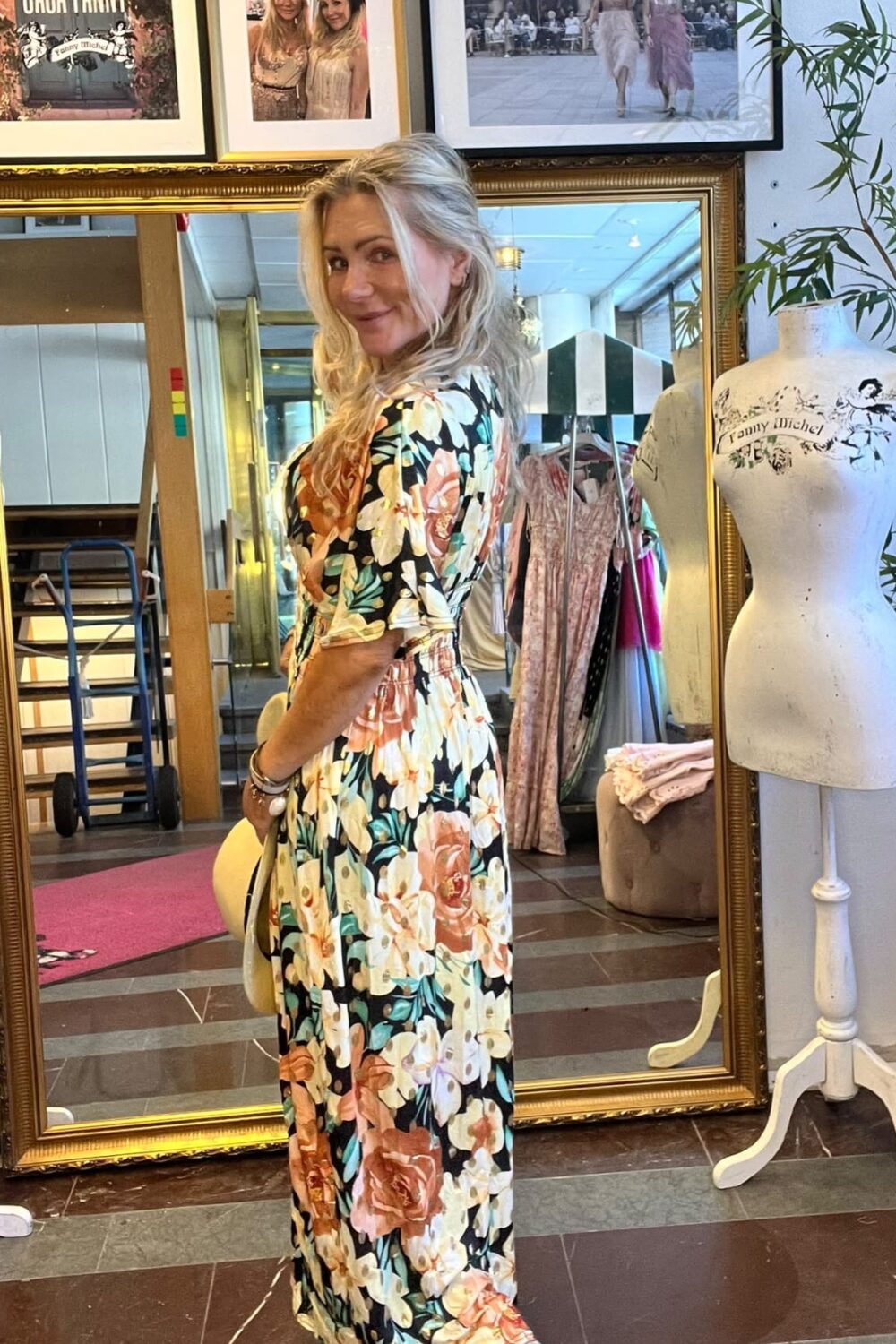 Blond kvinna poserar och ler i en blommig långklänning framför en stor guldramsspegel i en butik/ateljé. Runtom syns klädställning och provdocka med texten ”Fanny Michel” samt inramade foton ovanför.