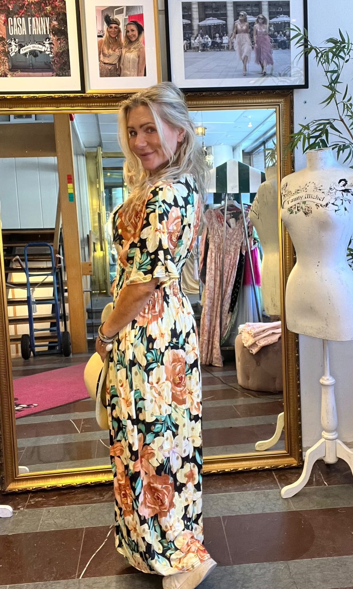 Blond kvinna poserar och ler i en blommig långklänning framför en stor guldramsspegel i en butik/ateljé. Runtom syns klädställning och provdocka med texten ”Fanny Michel” samt inramade foton ovanför.