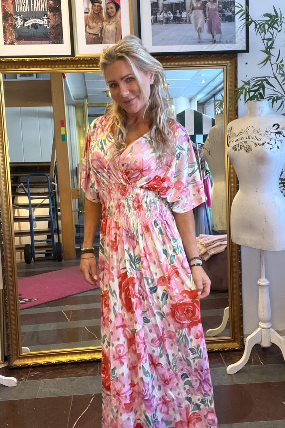 Kvinna står och ler mot kameran i en butik eller ateljé, iklädd en rosa blommig maxiklänning. Bakom henne syns en stor guldramsspegel, inramade foton med texten ”CASA FANNY”, och en provdocka.