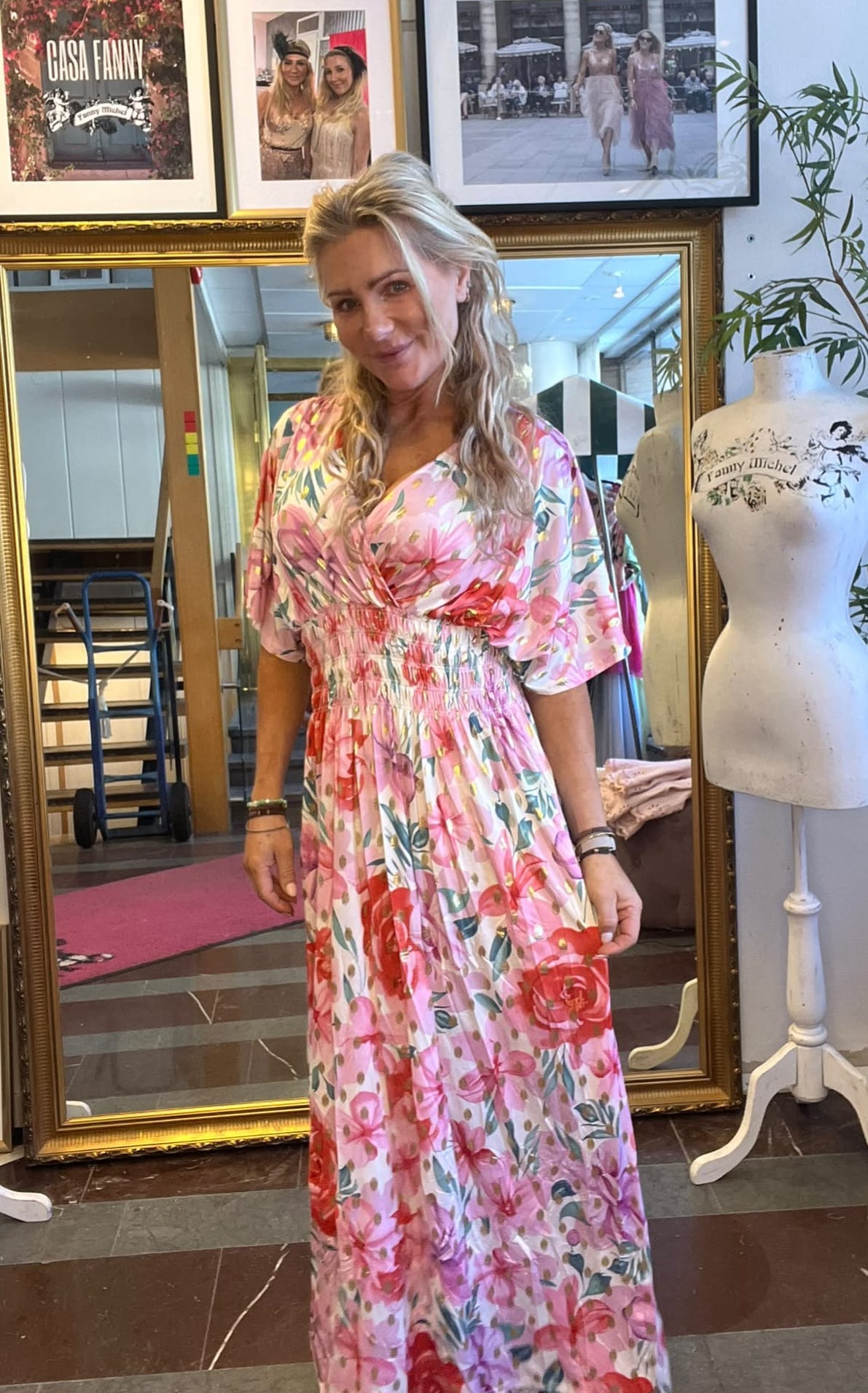 Kvinna står och ler mot kameran i en butik eller ateljé, iklädd en rosa blommig maxiklänning. Bakom henne syns en stor guldramsspegel, inramade foton med texten ”CASA FANNY”, och en provdocka.