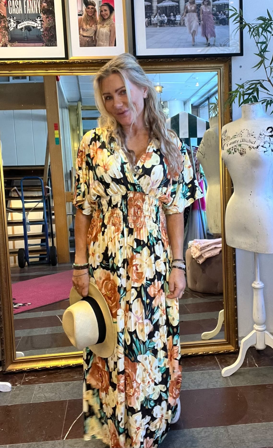 Blond kvinna poserar leende i lång blommig klänning och håller en ljus stråhatt med svart band. Hon står i en butik framför stor guldramsspegel; bredvid syns provdocka och inramade foton med texten “CASA FANNY” och “Fanny Michel”.