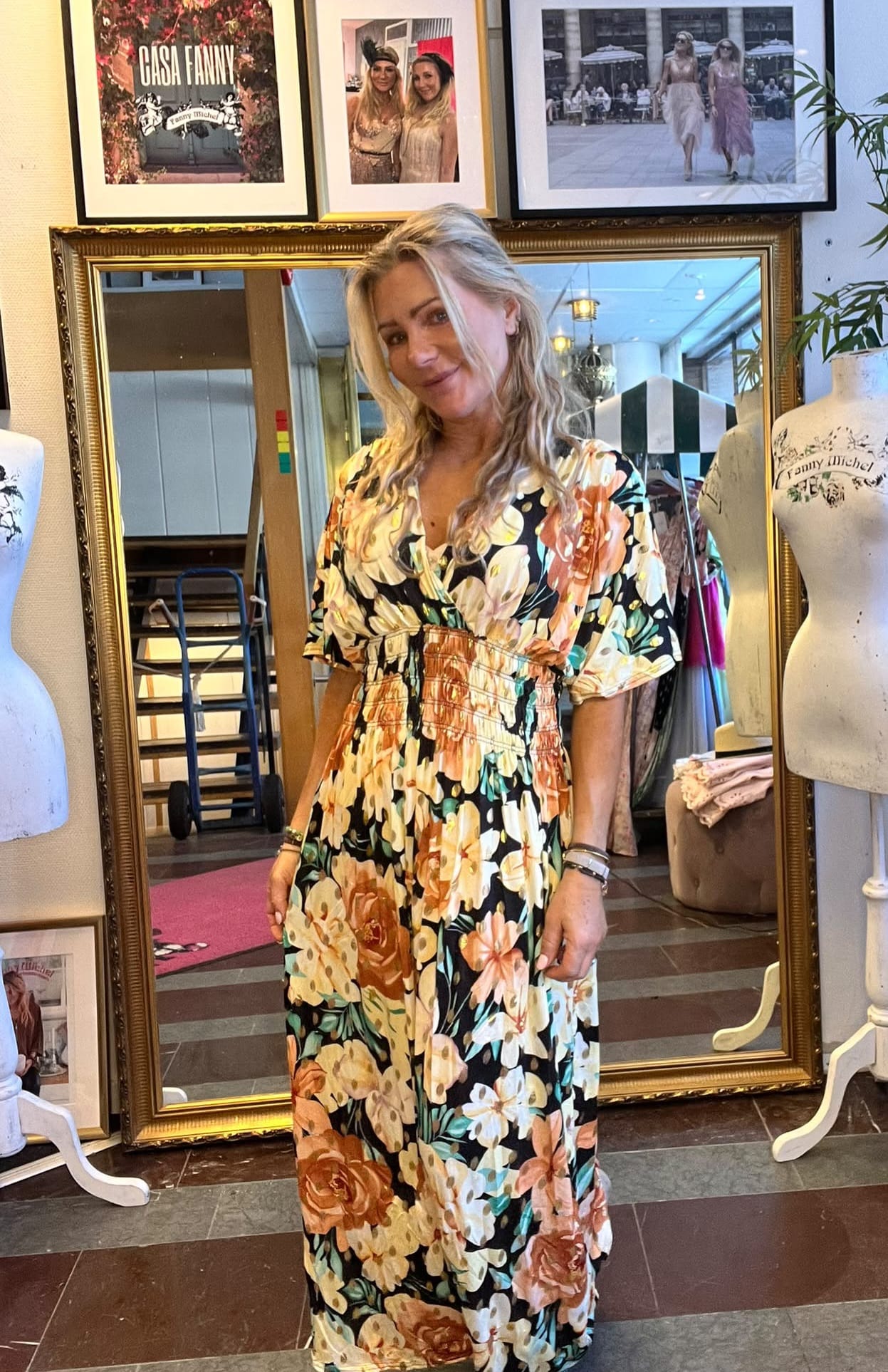 Blond kvinna står och ler framför en stor guldramsspegel i en klädbutik, iklädd lång blommig klänning. Omgiven av provdockor och klädställningar; på väggen ovanför syns inramade bilder med texten “CASA FANNY”.