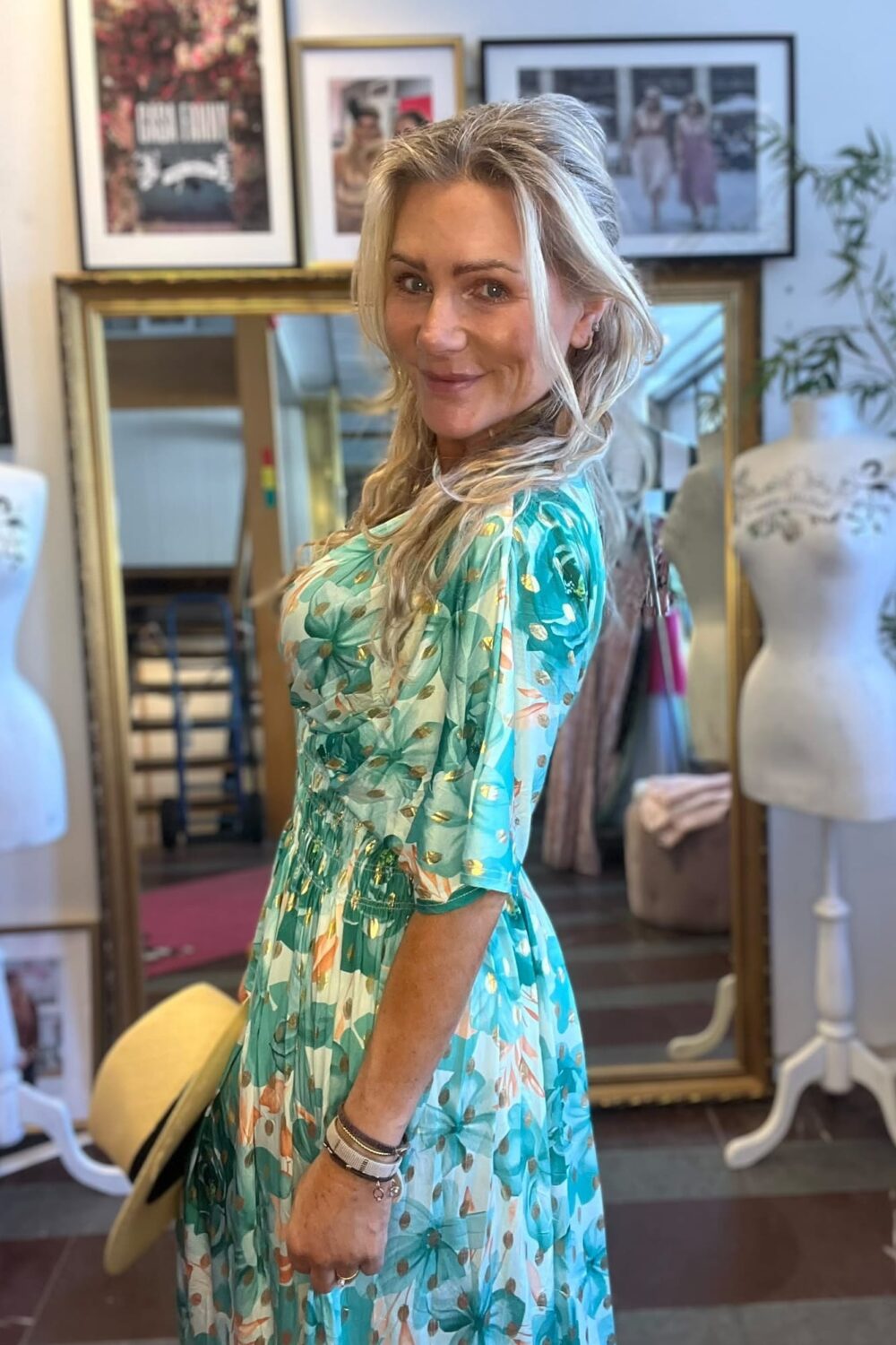 Blond kvinna i grönmönstrad klänning poserar och ler mot kameran med en ljus hatt i handen. Hon står i en butik eller ateljé med speglar, provdockor och inramade foton i bakgrunden.