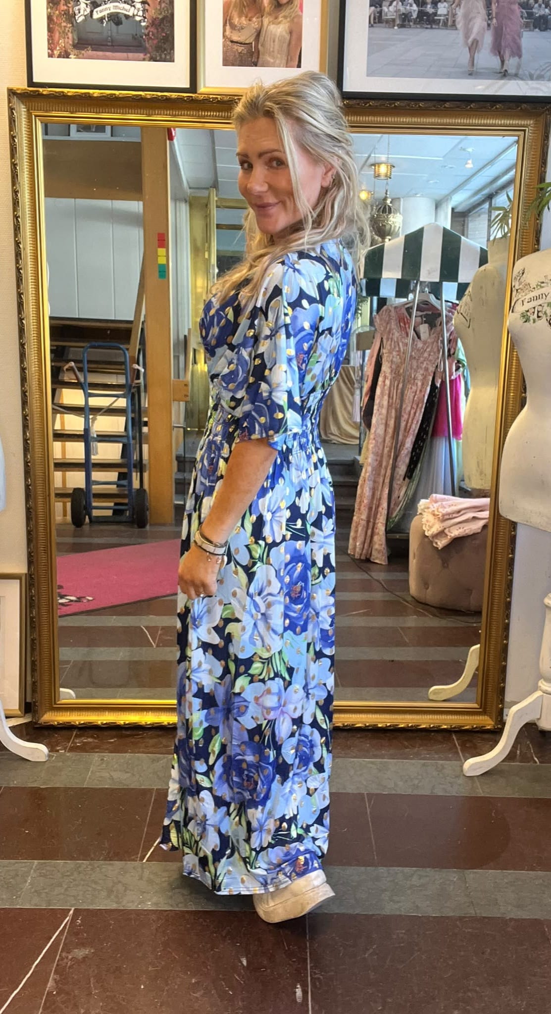 Kvinna i blå, lång blommig klänning står i profil och ler mot kameran framför en stor guldramsspegel i en butik, med klädställning, provdocka och inramade foton på väggen bakom.