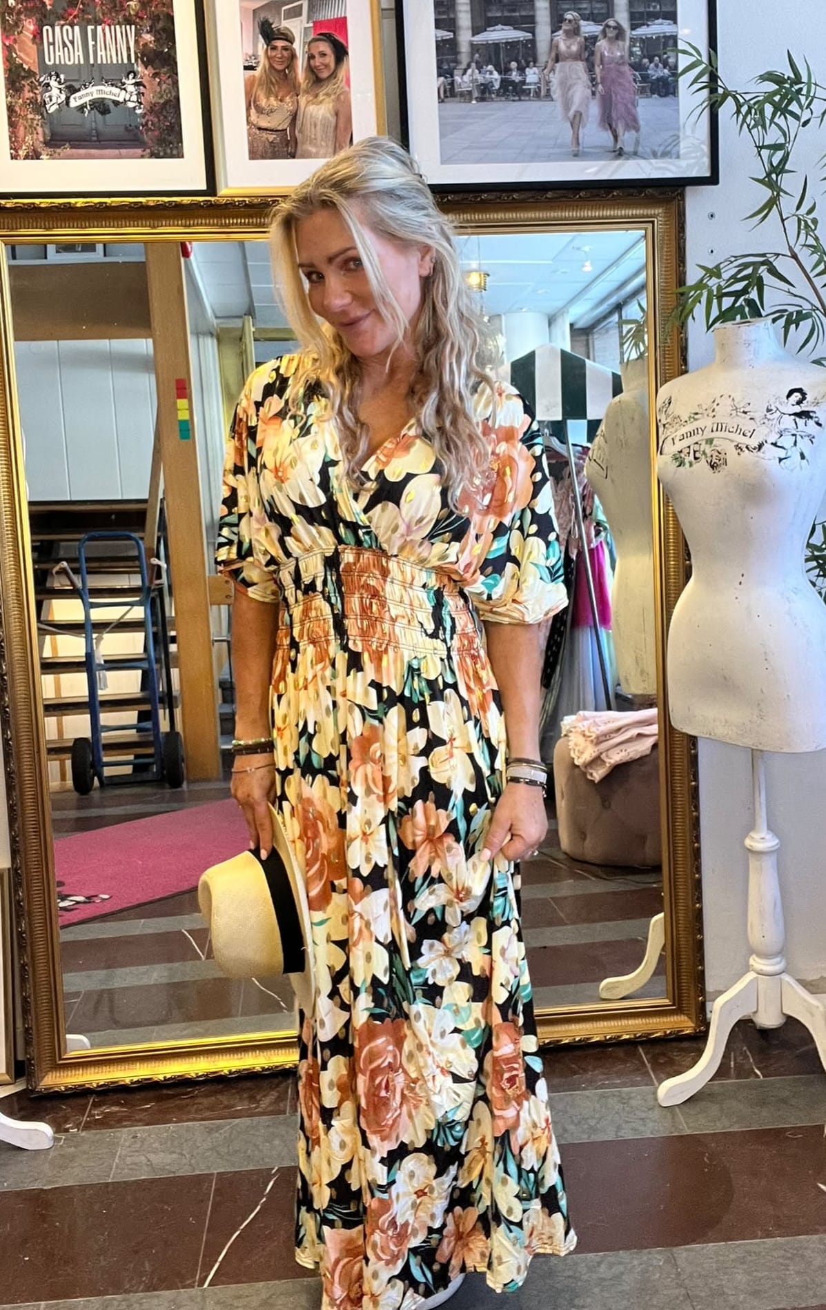 Blond kvinna poserar och håller en halmhatt vid sidan, klädd i lång blommig klänning, i en boutique/ateljé. Bakom syns stor guldspegel, skyltdocka med texten ”Fanny Michel” samt inramade foton och ”CASA FANNY”.