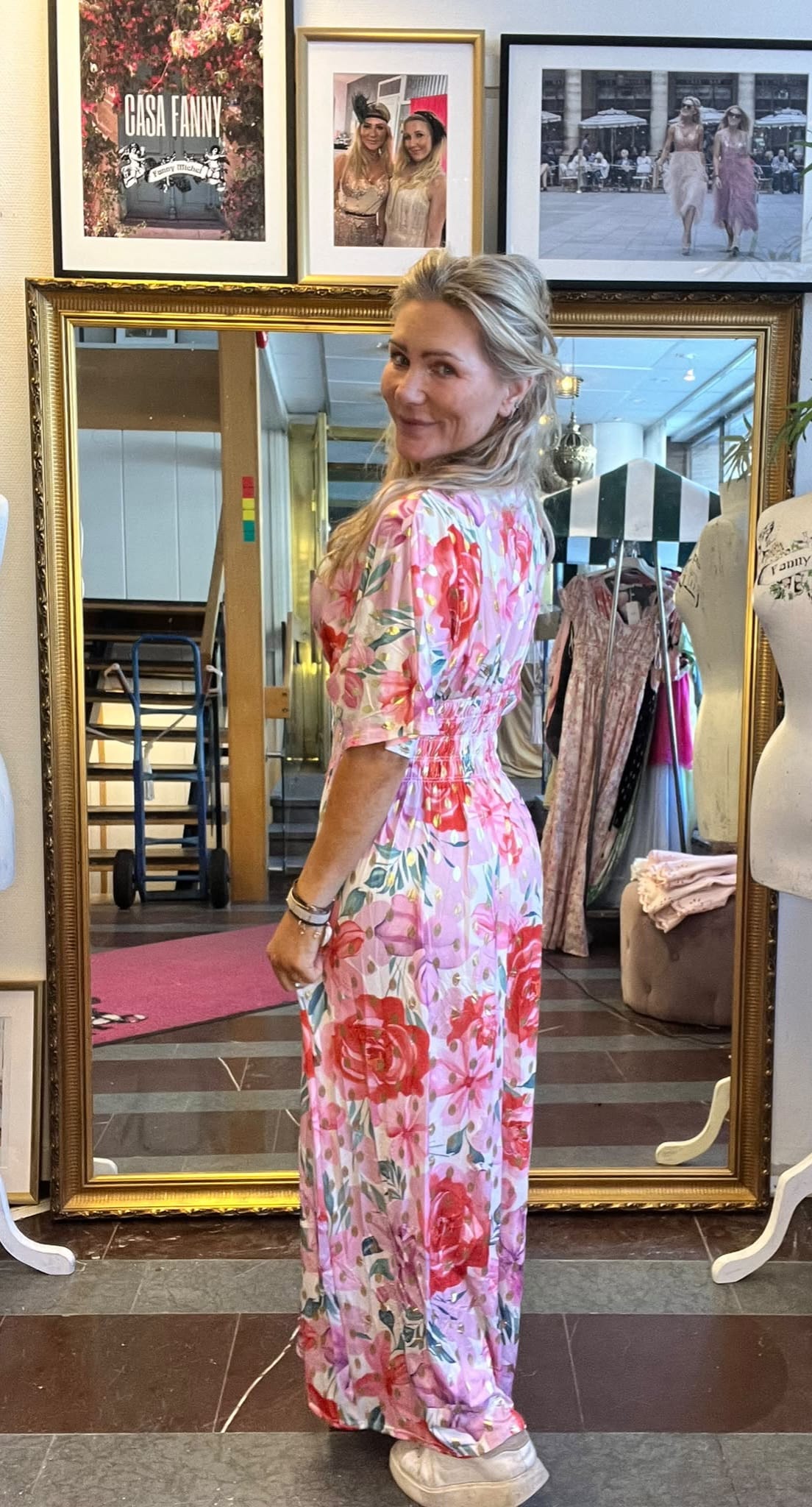 Kvinna står framför en stor guldramsspegel och vänder sig leende mot kameran, iklädd lång blommig klänning i rosa och rött. Runt spegeln syns klädställning och provdocka; ovanför hänger ramar med texten “CASA FANNY”.