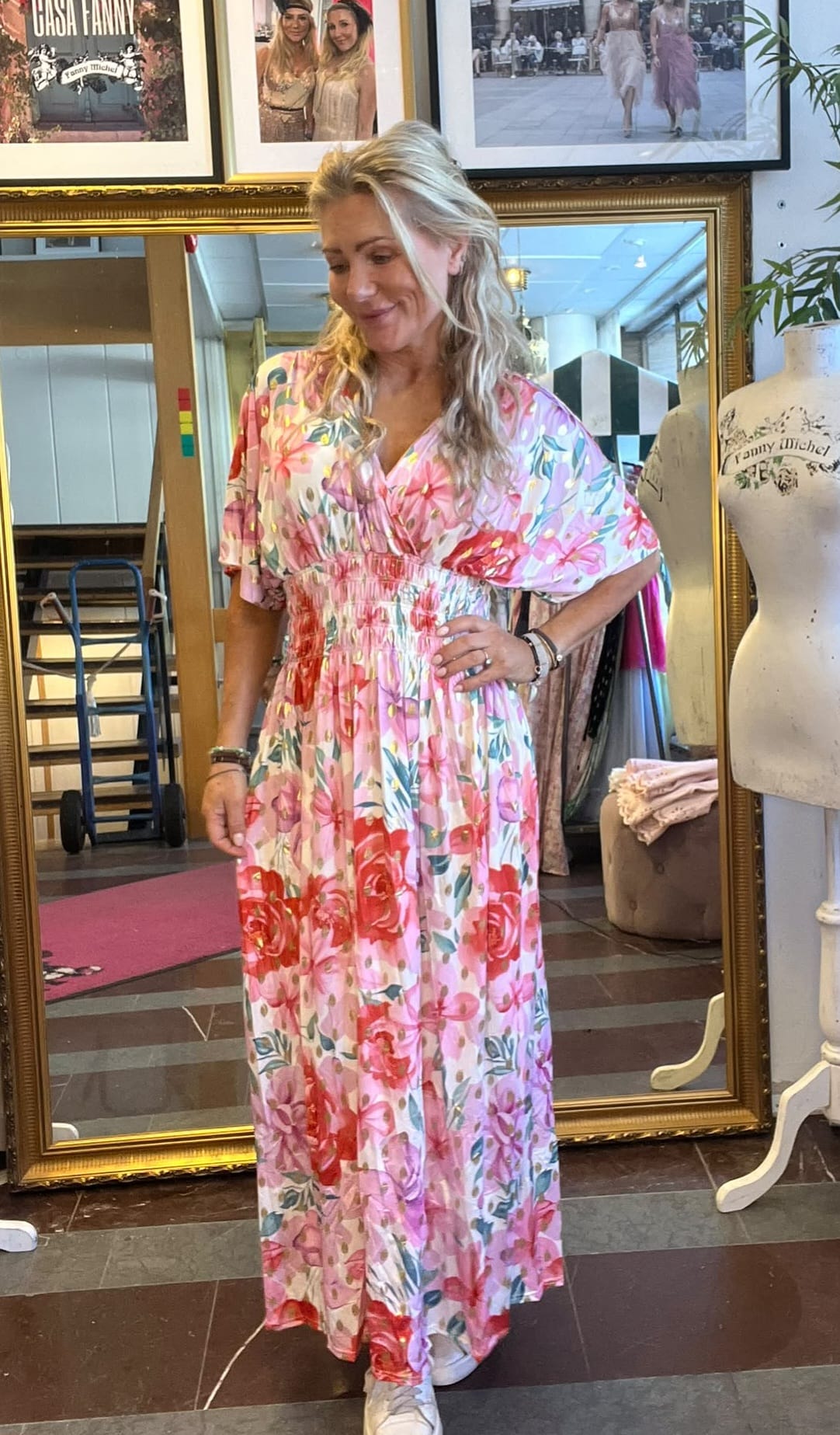 Blond kvinna provar en lång, rosa blommig klänning och ser ner med handen på höften i en butik. Bakom henne står en stor spegel, skyltdocka med texten “Fanny Michel” och inramade foton på väggen.