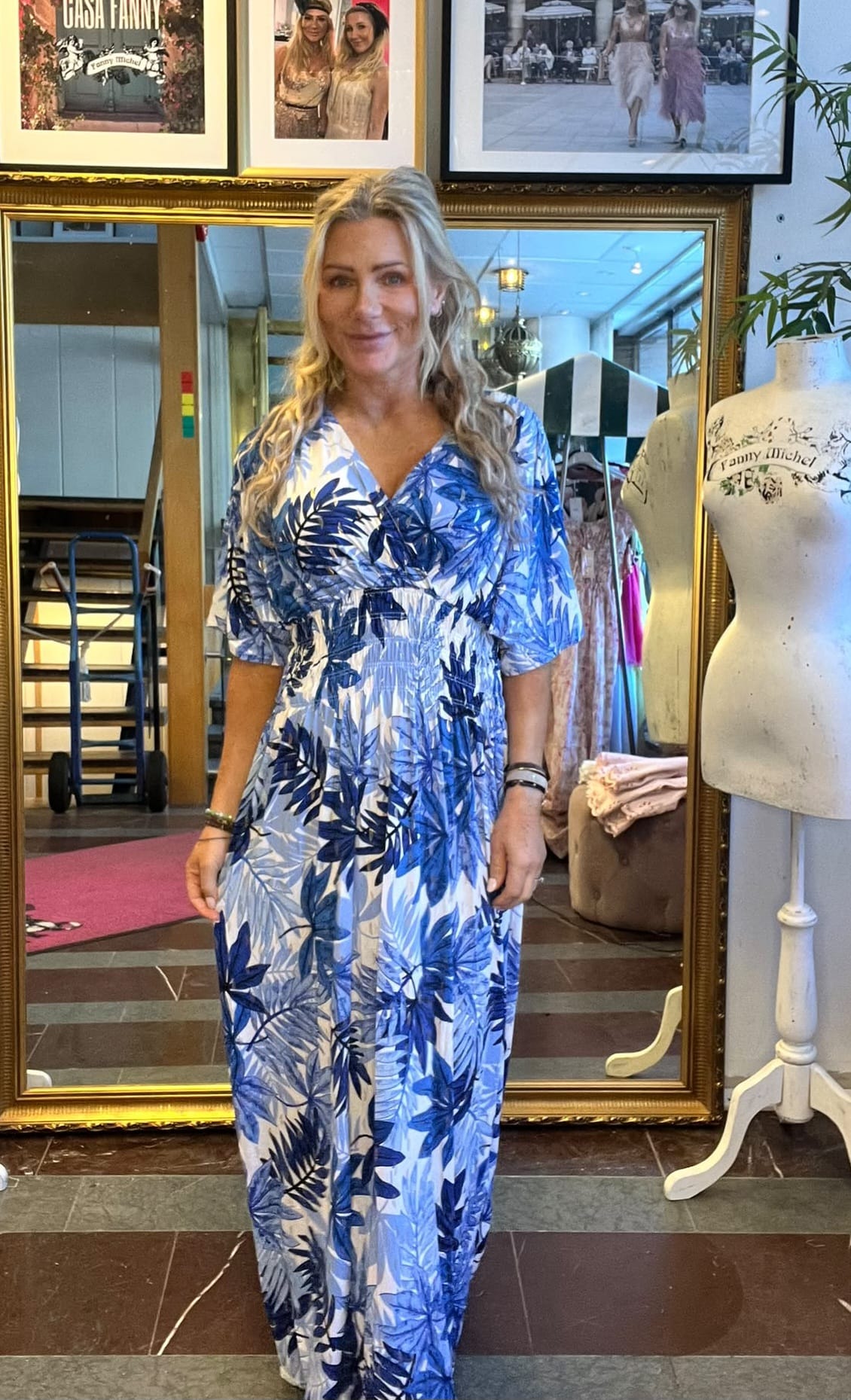 Leende kvinna står framför en stor guldramsspegel och visar upp en lång blåvit klänning med bladmönster i en butik. Bredvid syns en provdocka med texten “Fanny Michel” och inramade foton på väggen.
