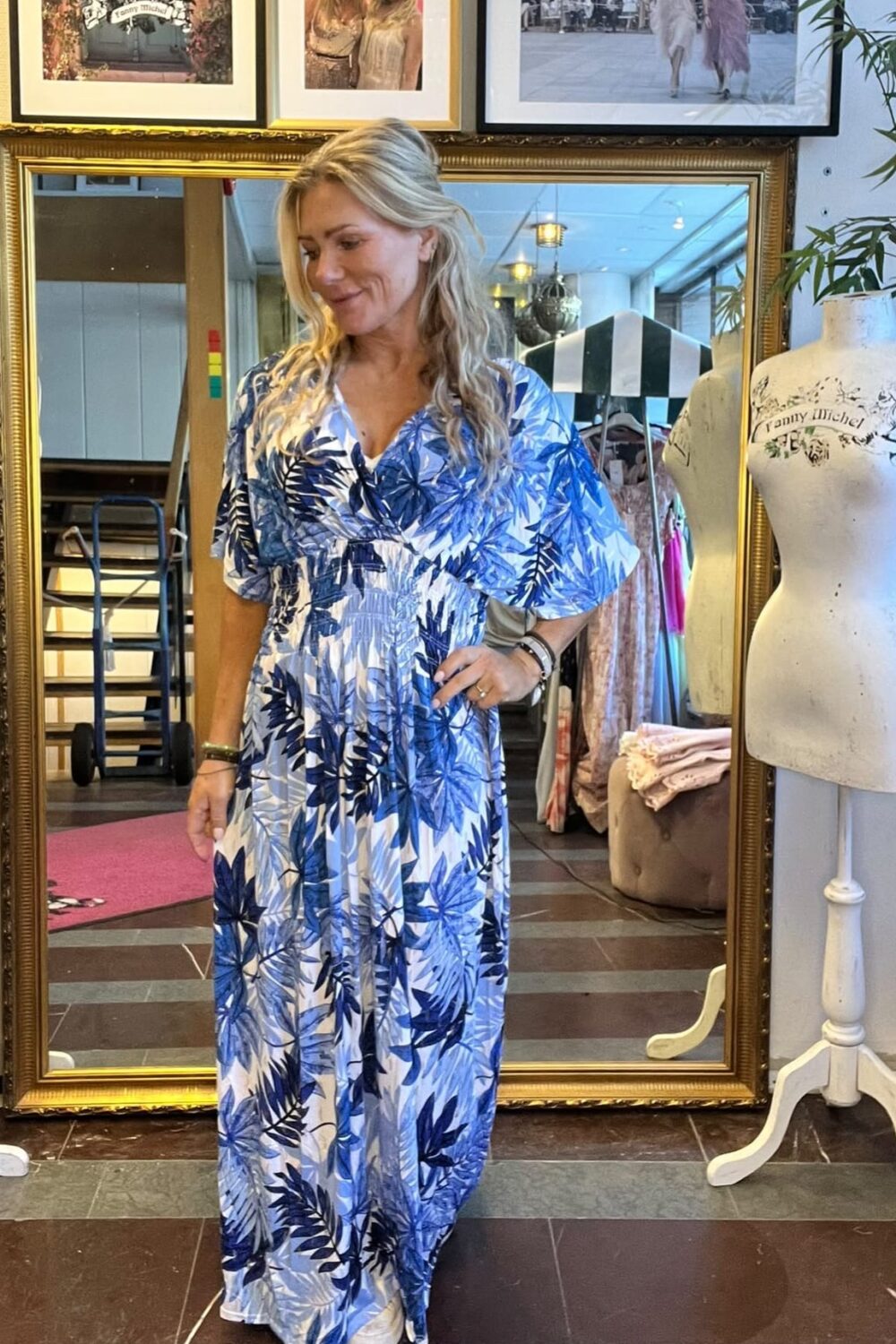Kvinna med långt blont hår provar en blåvit långklänning med bladmönster och ser ned med ett leende. Hon står framför en stor guldramad spegel i en butik, bredvid en provdocka med texten “Fanny Michel”.