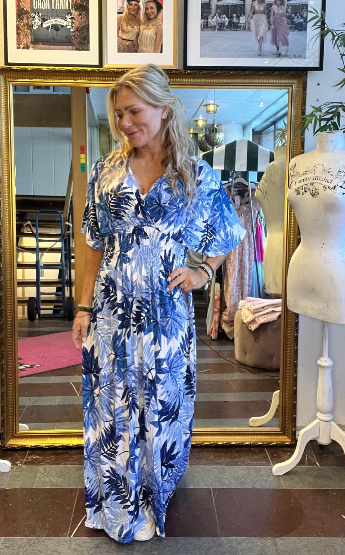 Kvinna med långt blont hår provar en blåvit långklänning med bladmönster och ser ned med ett leende. Hon står framför en stor guldramad spegel i en butik, bredvid en provdocka med texten “Fanny Michel”.