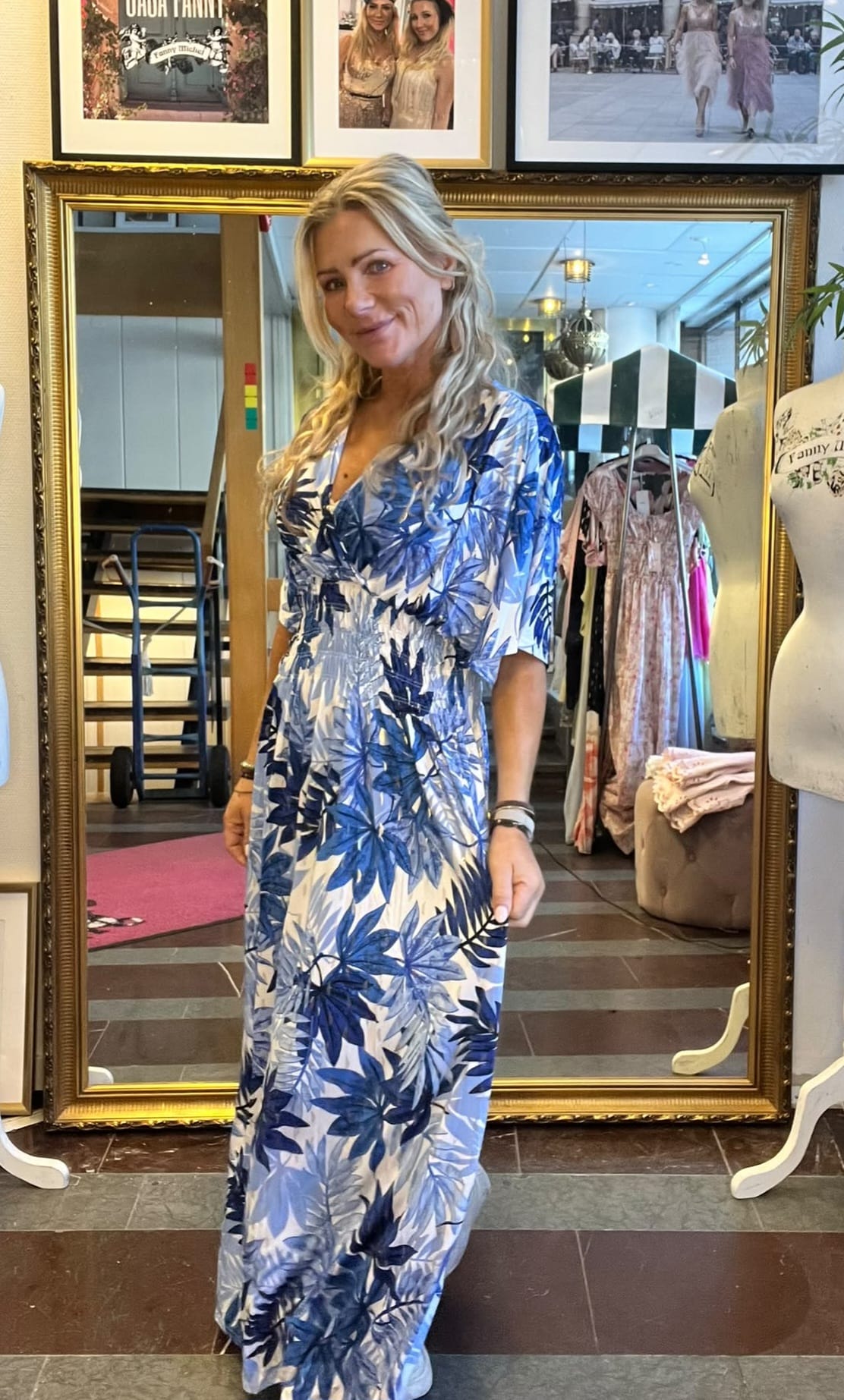 Blond kvinna står och poserar i en klädbutik, iklädd en lång blåvit blommig klänning och håller ut tyget med handen. Bakom henne syns en stor guldramsspegel, klädställning, provdocka och inramade foton.