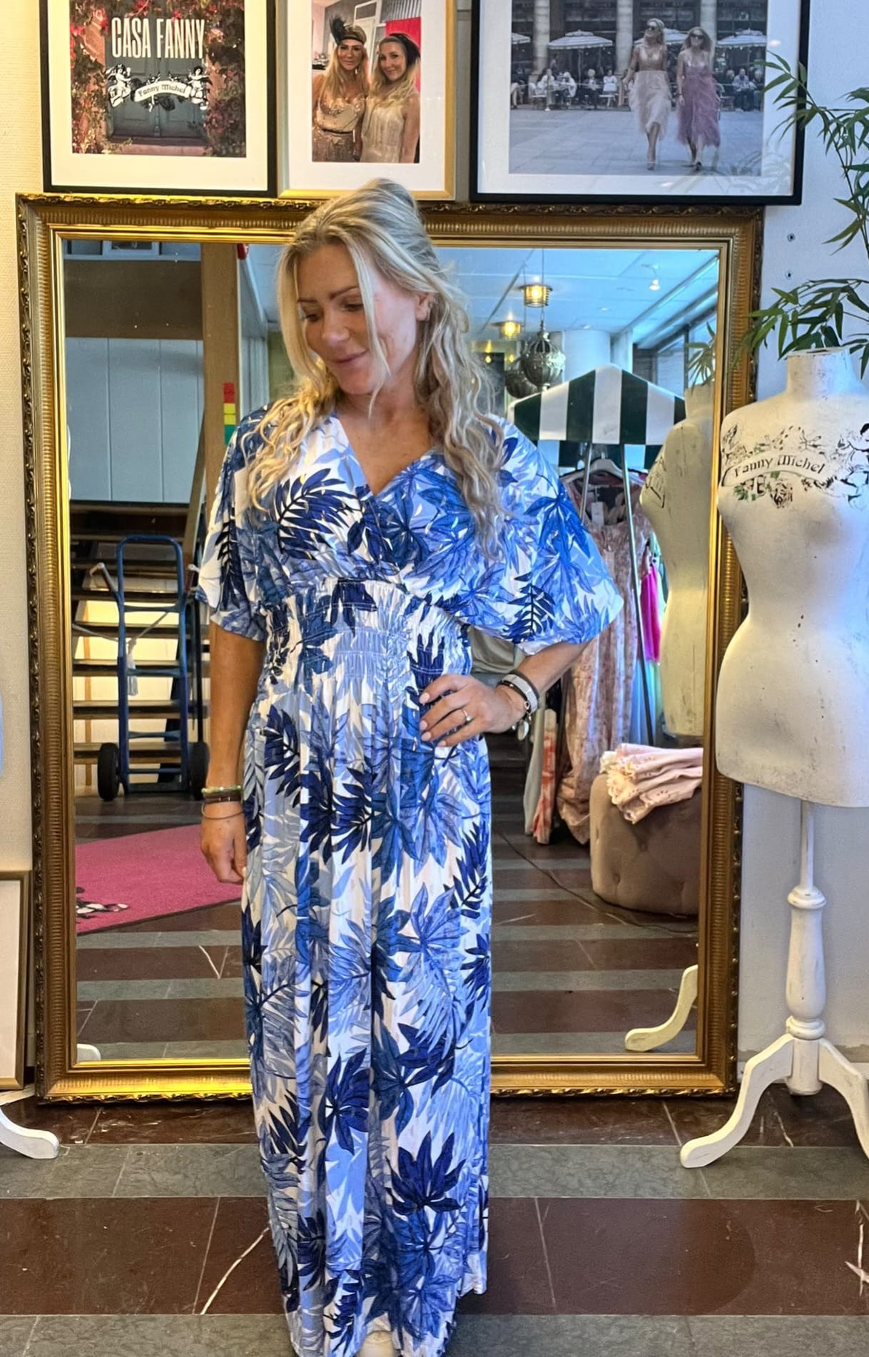 Kvinna provar en blåvit, blommönstrad långklänning och poserar med handen på höften framför en stor guldspegel i en butik. Intill står en provdocka med texten “Fanny Michel”, och ovanför hänger inramade foton.