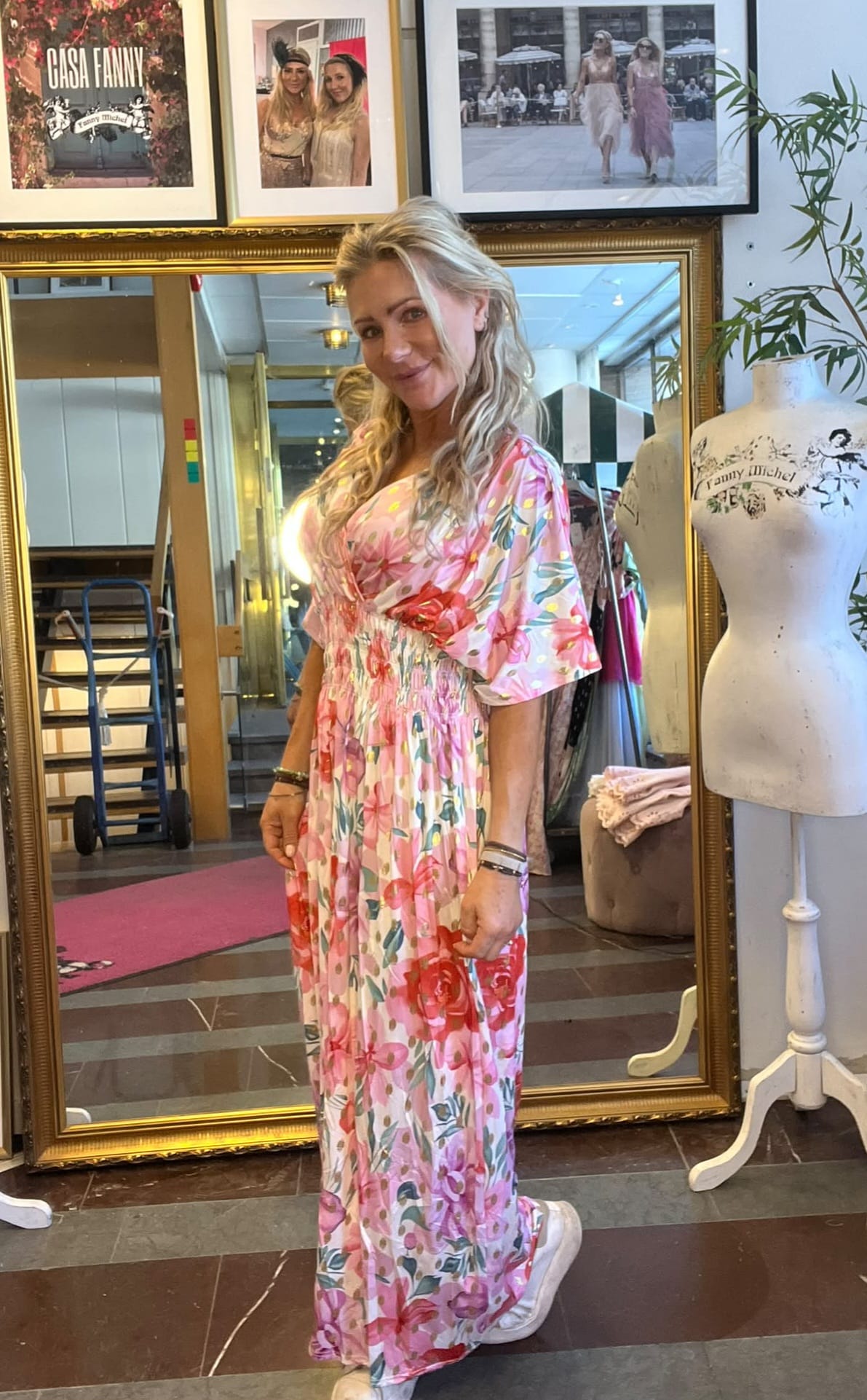 Kvinna i lång rosa blommig klänning står och ler framför en stor guldramsspegel i en klädbutik. I bakgrunden syns provdocka, klädställning och inramade foton med texten “CASA FANNY” och “Fanny Michel”.