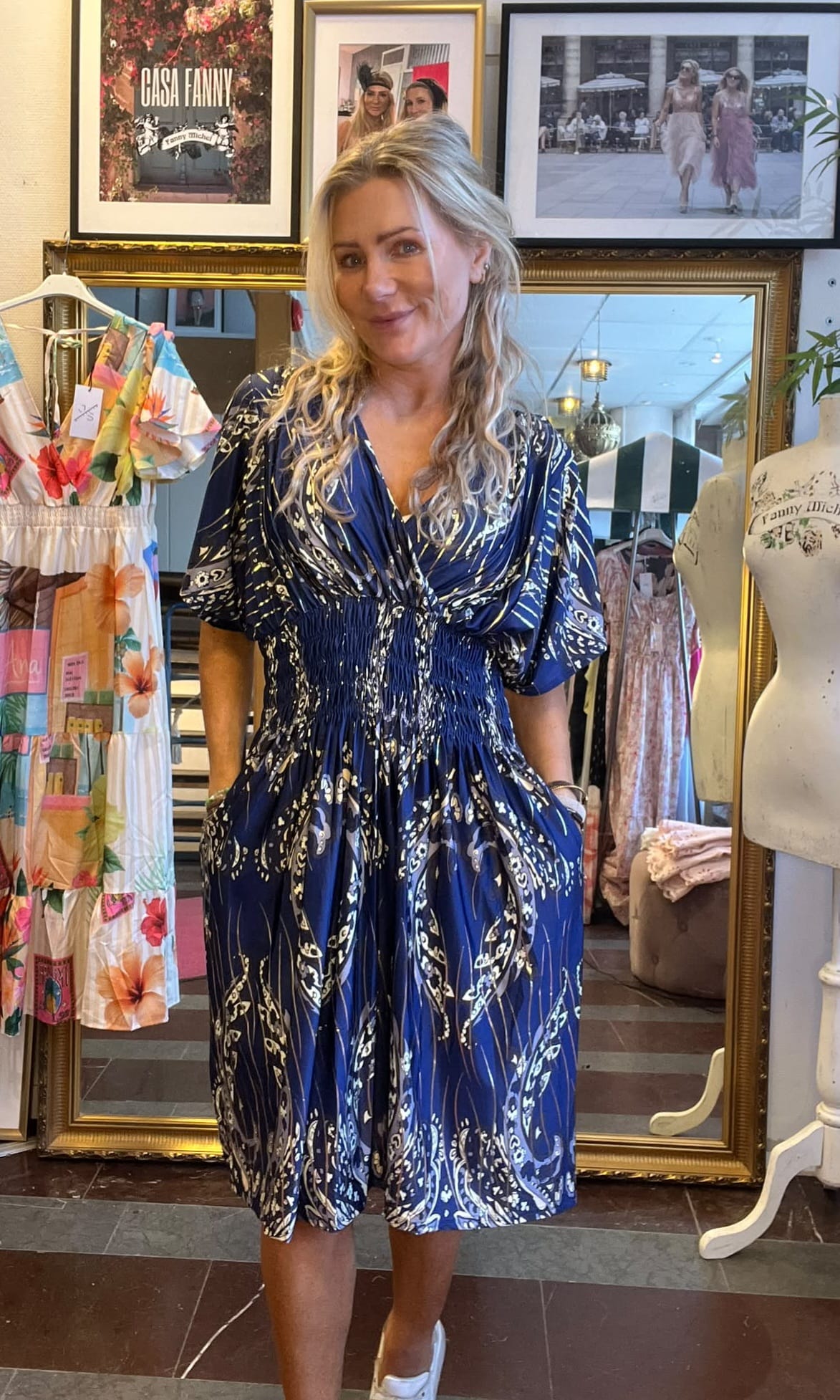 Blond kvinna står och ler med händerna i fickorna, klädd i en blå mönstrad klänning, i en klädbutik. Bakom henne syns klänningar på galge, provdocka och inramade foton med texten “CASA FANNY”.