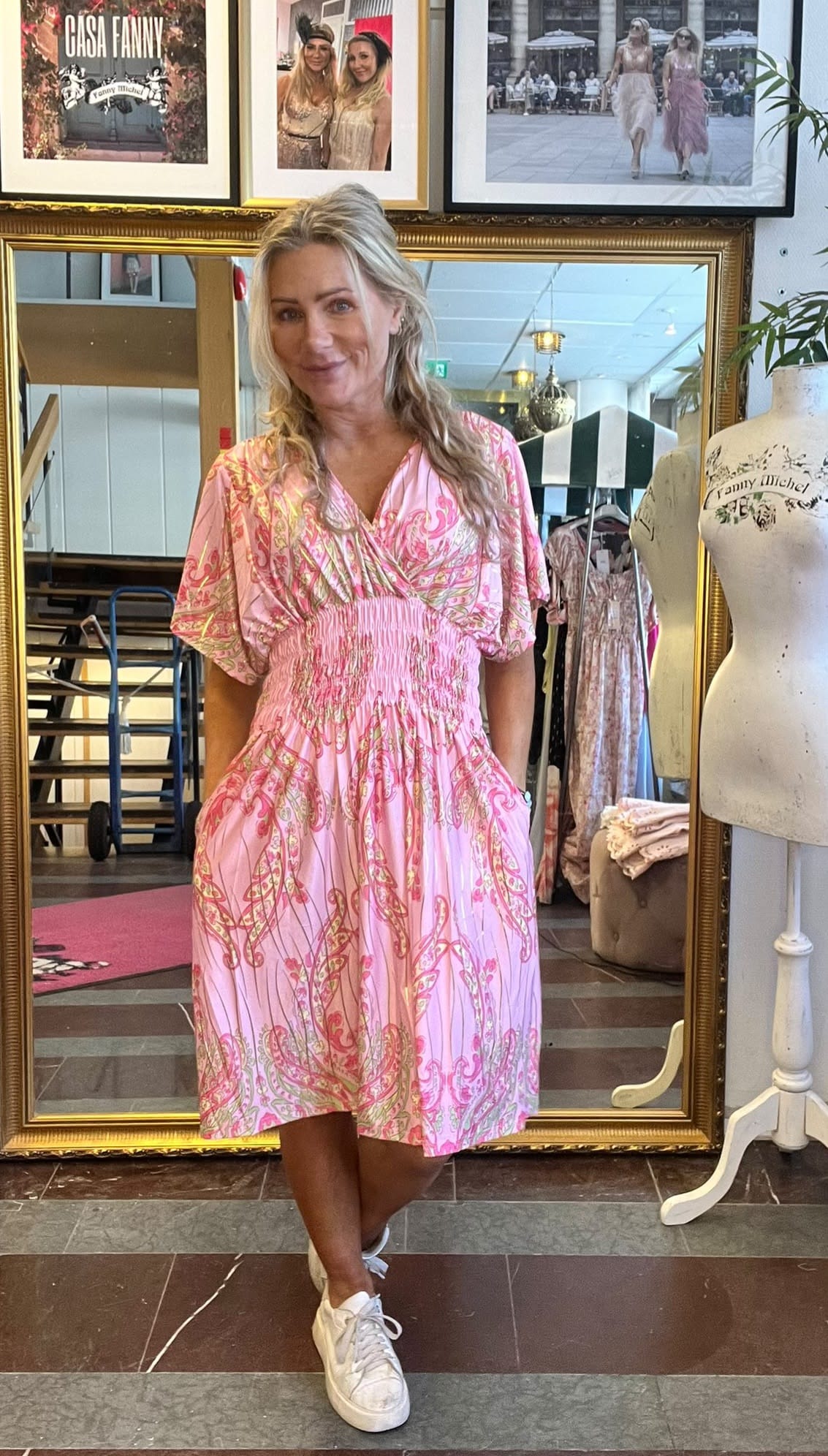 Blond kvinna står och ler med händerna i fickorna, iklädd rosa mönstrad klänning och vita sneakers. Hon poserar framför en stor guldspegel i en butik/ateljé med skyltdocka och inramade foton; text: “CASA FANNY”.