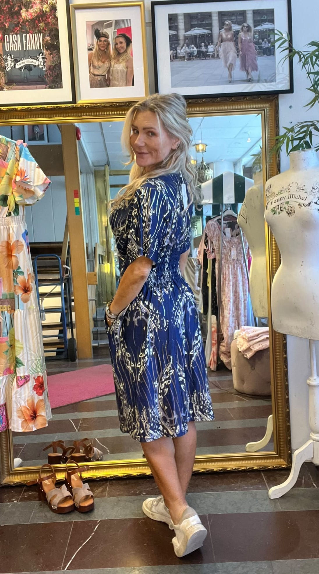 Blond kvinna provar en blå, mönstrad klänning och ler över axeln framför en stor guldspegel i en klädbutik. Runt henne syns klädställningar, sandaler på golvet och en provdocka med texten “Fanny Michel”.