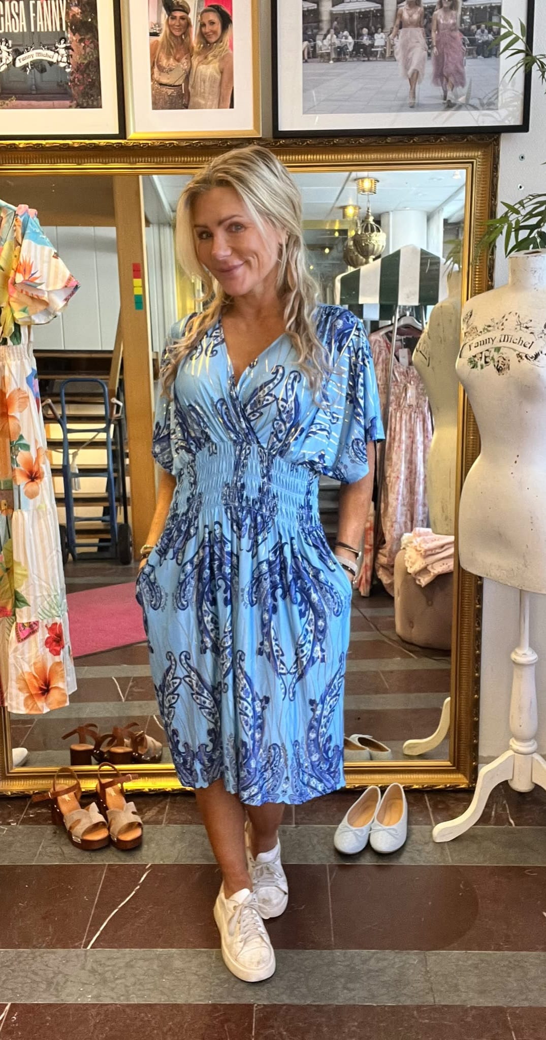 Blond kvinna står och ler framför en stor guldspegel i en klädbutik, med händerna i fickorna på en ljusblå mönstrad klänning och vita sneakers. Runtom syns skor, klänningar och provdocka med texten “Fanny Michel”.