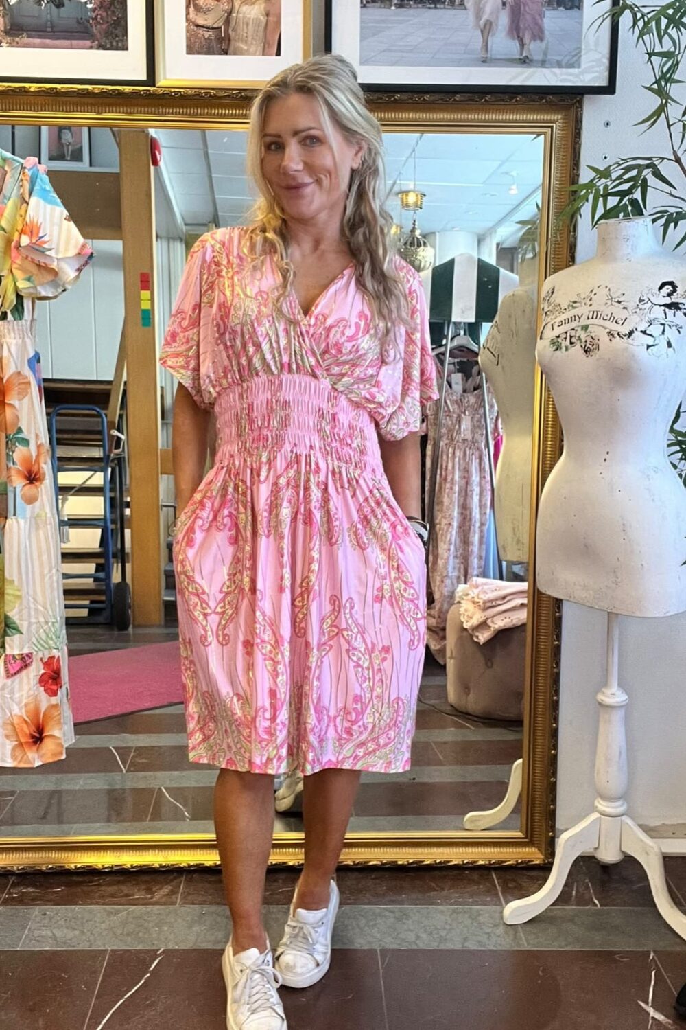 Kvinna står och ler i en klädbutik, klädd i rosa mönstrad klänning och vita sneakers. Bakom henne syns en stor guldramsspegel, upphängda klänningar och fotokonst; provdocka med texten “Fanny Michel”.