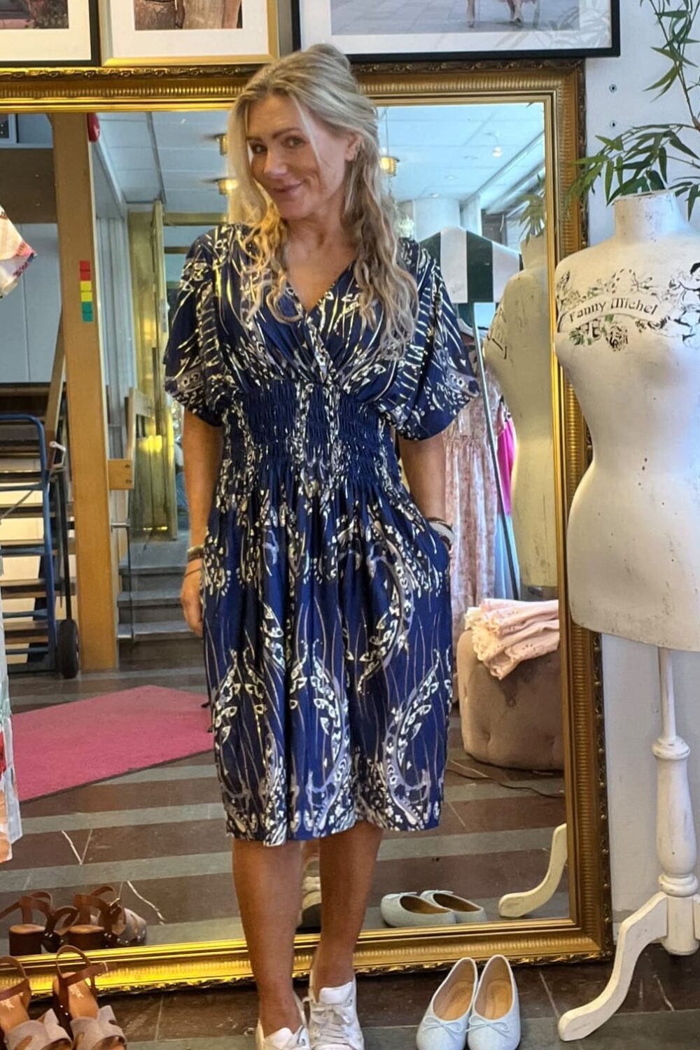 Blond kvinna står och ler framför en stor guldramsspegel i en klädbutik, klädd i blå mönstrad klänning och vita sneakers. Runt henne syns skor på golvet, klädställning och en provdocka med texten ”Fanny Michel”.
